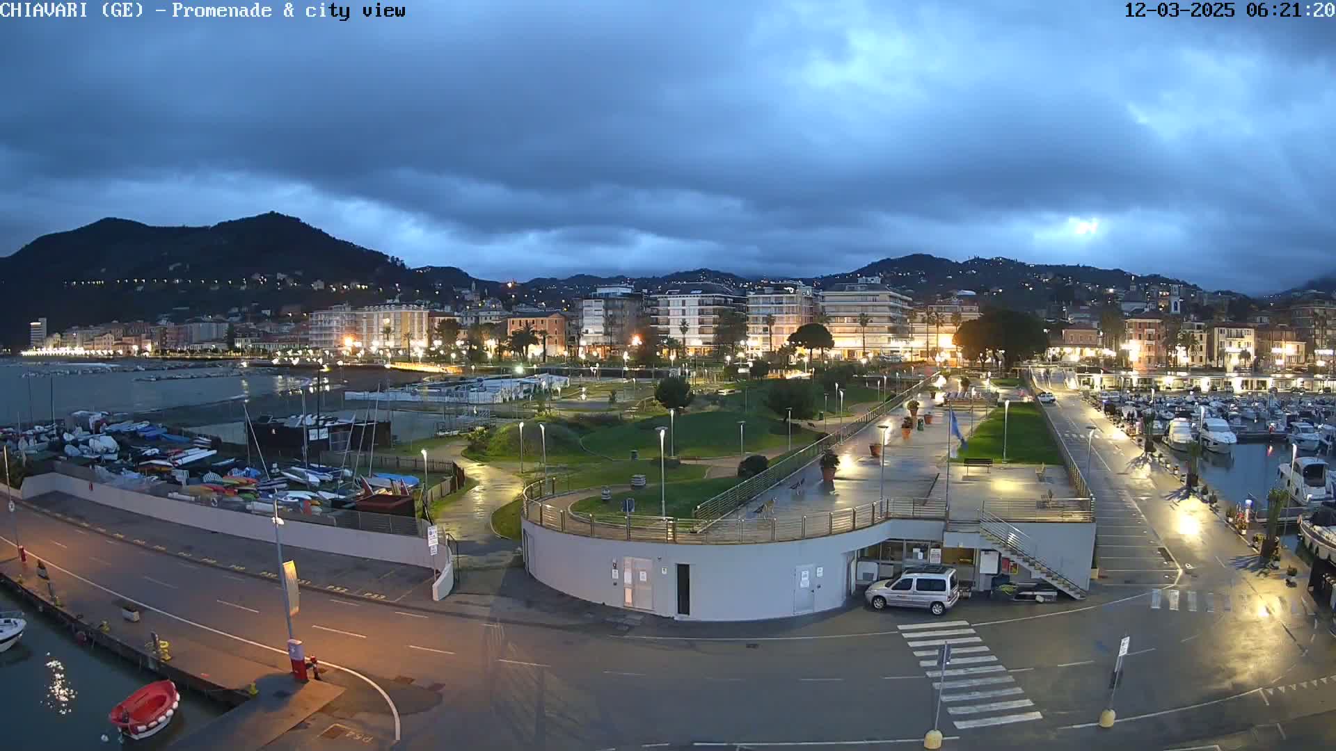A city's waterfront at twilight under a cloudy sky shows a marina filled with boats, a promenade with lights, and buildings illuminated against a hill.