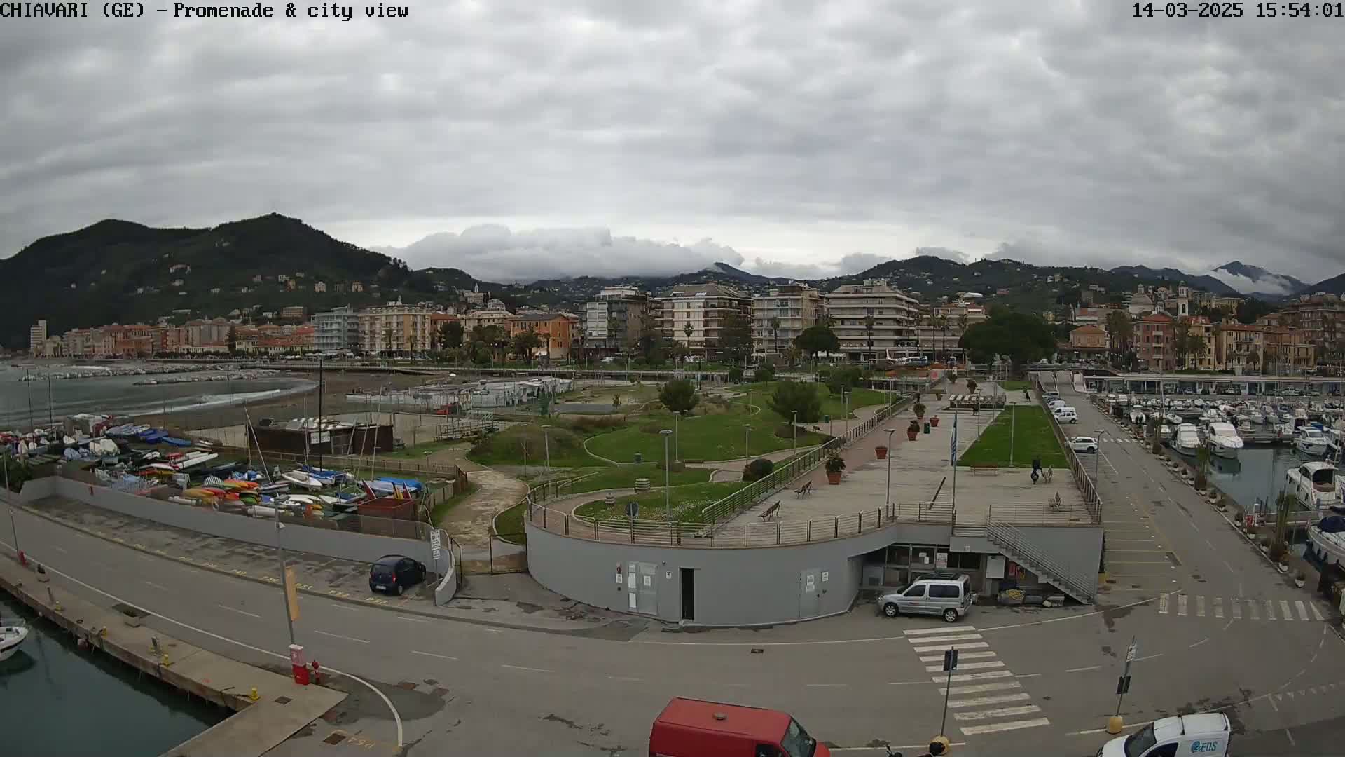 An overcast day reveals a coastal city with a marina, promenade, and park, backed by mountains partially shrouded in clouds.
