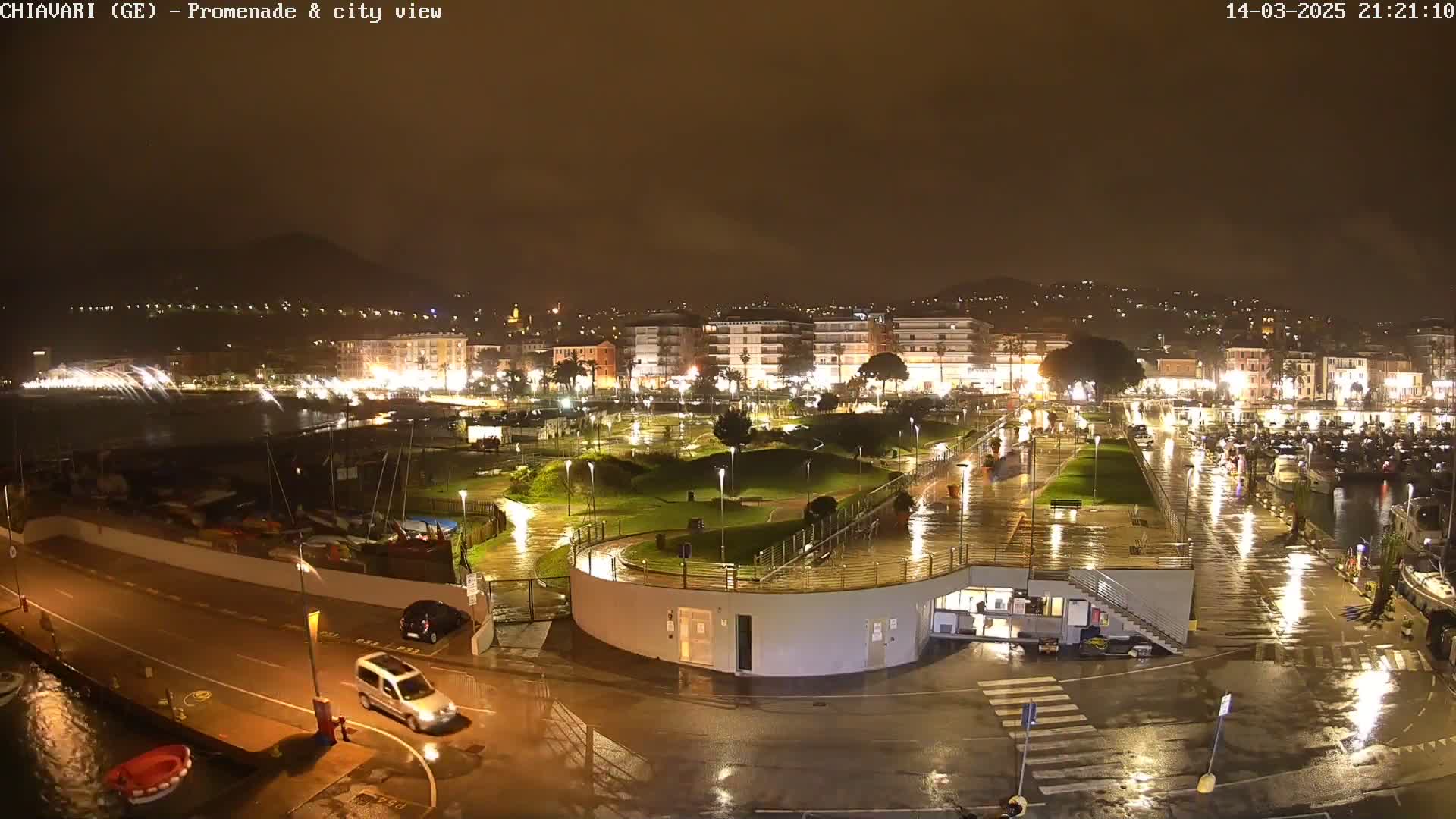 A rain-slicked, nighttime view of a city's waterfront promenade and harbor, with illuminated buildings and parked cars reflecting in the wet surfaces.