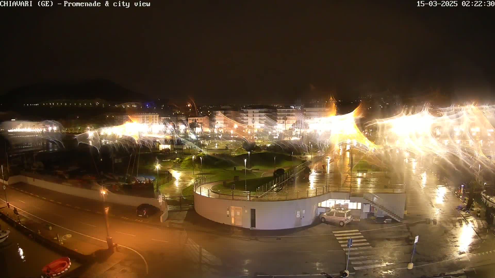 A nighttime view of a rain-soaked city promenade and harbor, illuminated by bright lights and featuring streaks of light possibly from long exposure photography or moving water features.