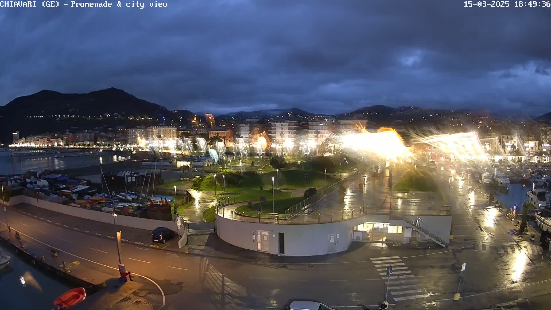 A nighttime view of a harbor town under a dark, cloudy sky, showing wet streets and a marina filled with boats, with bright lights illuminating a waterfront park.