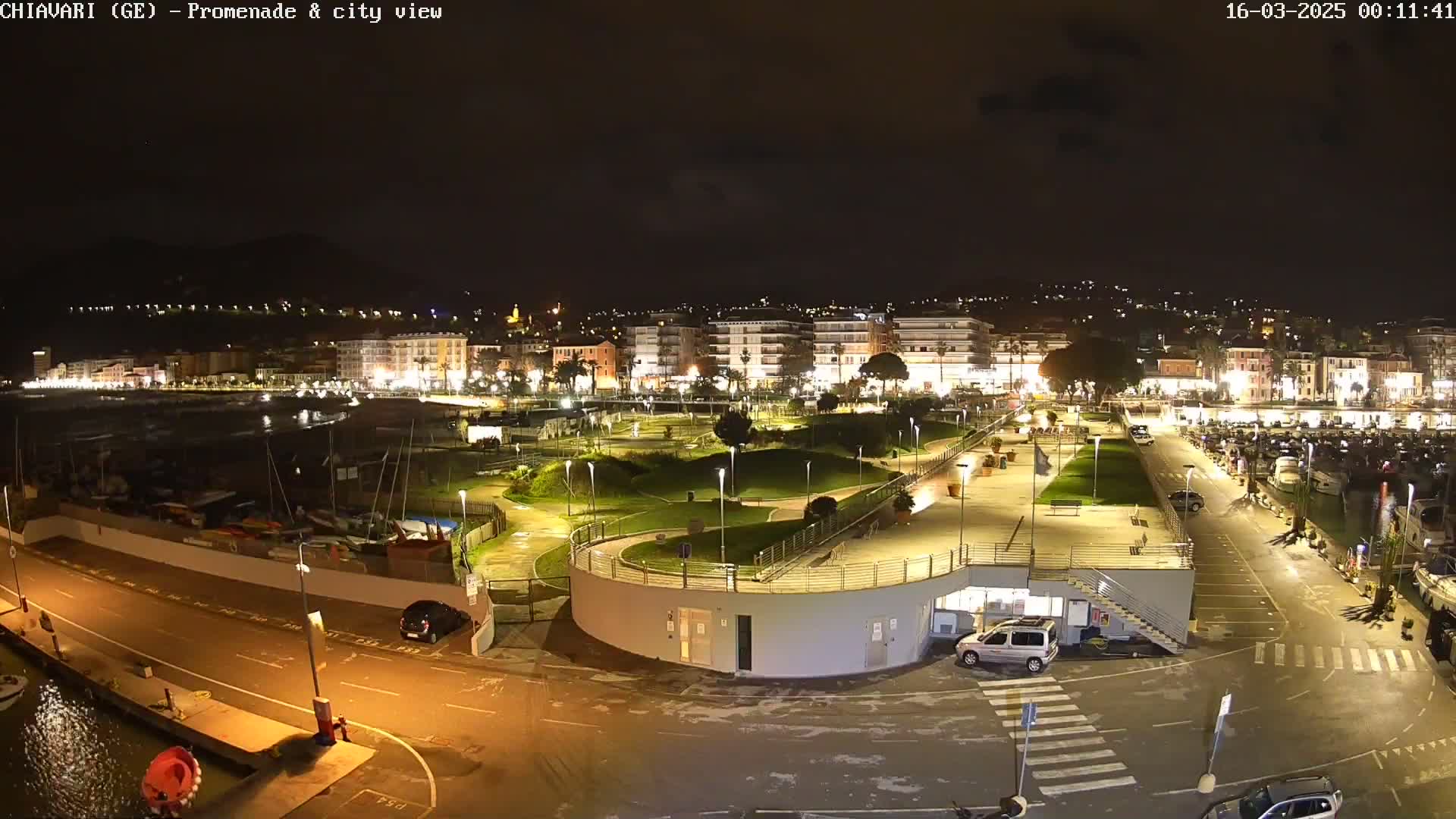 A nighttime view of a brightly lit waterfront promenade and city, with a marina containing several boats, and a parking lot with a few vehicles.