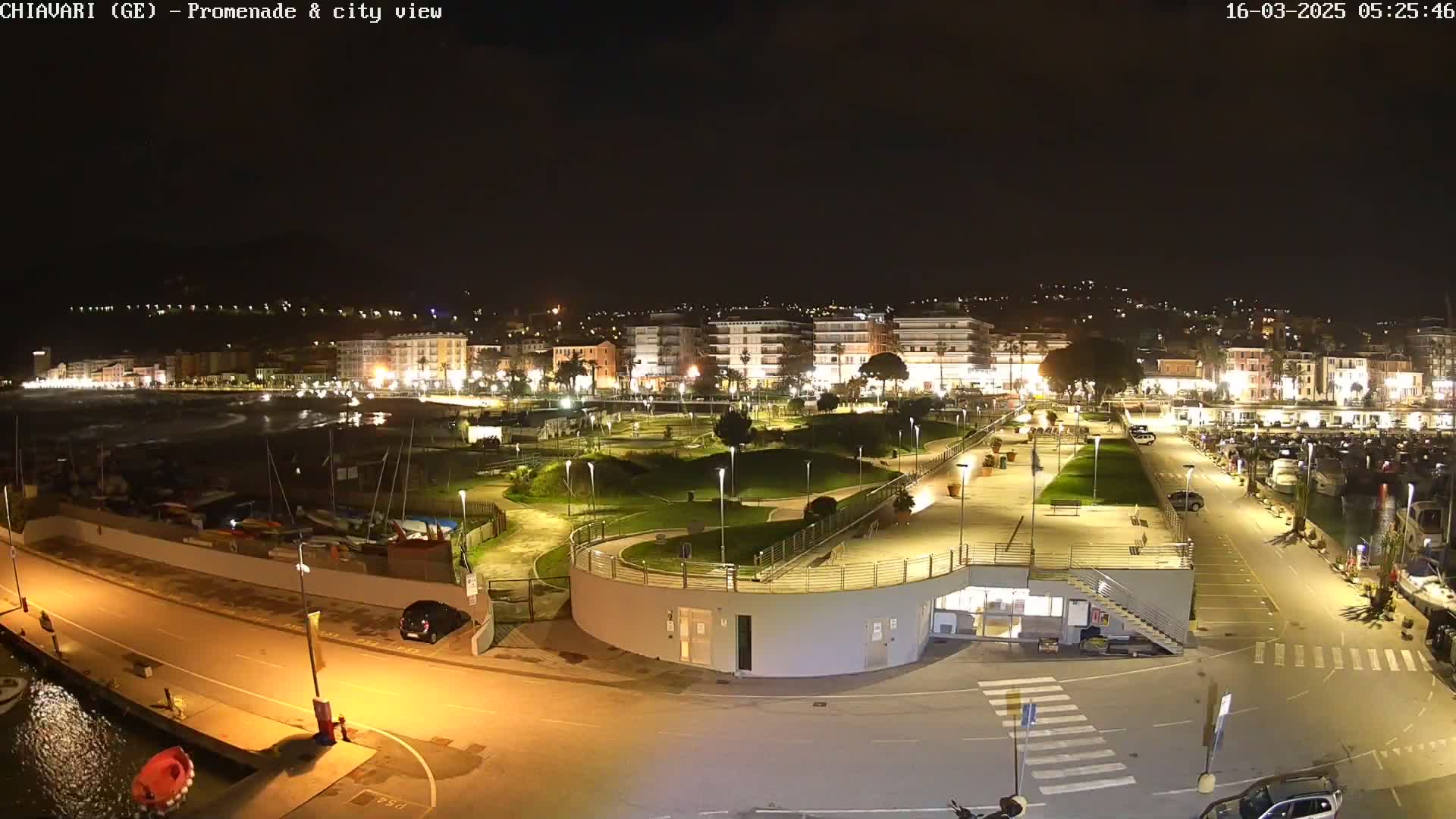 A nighttime, illuminated view of a waterfront promenade and city, with a calm body of water and boats visible in the distance.