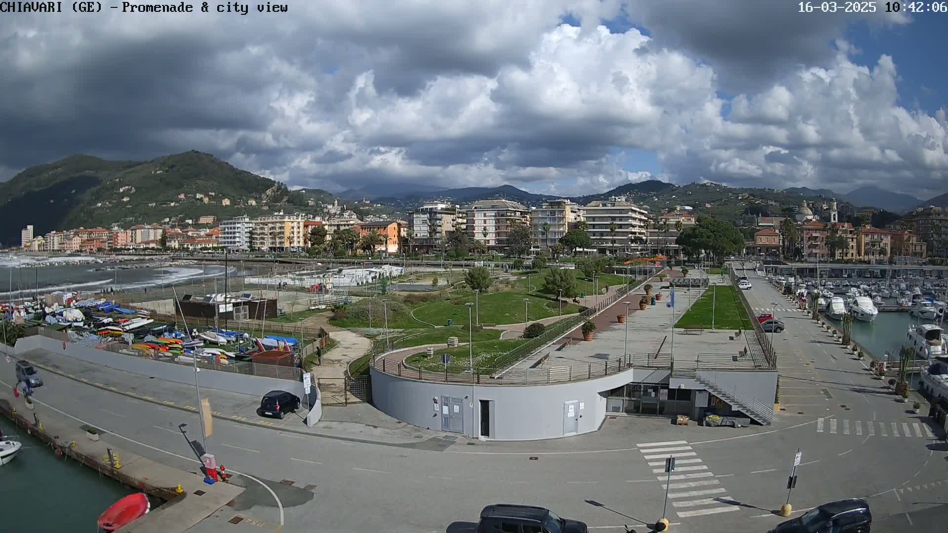 A partly cloudy day overlooks a coastal city with a promenade, marina, and a grassy park area.