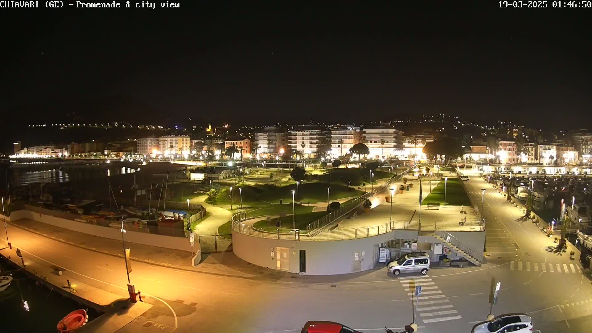A nighttime view of a waterfront promenade and city, with a well-lit park and harbor filled with boats.