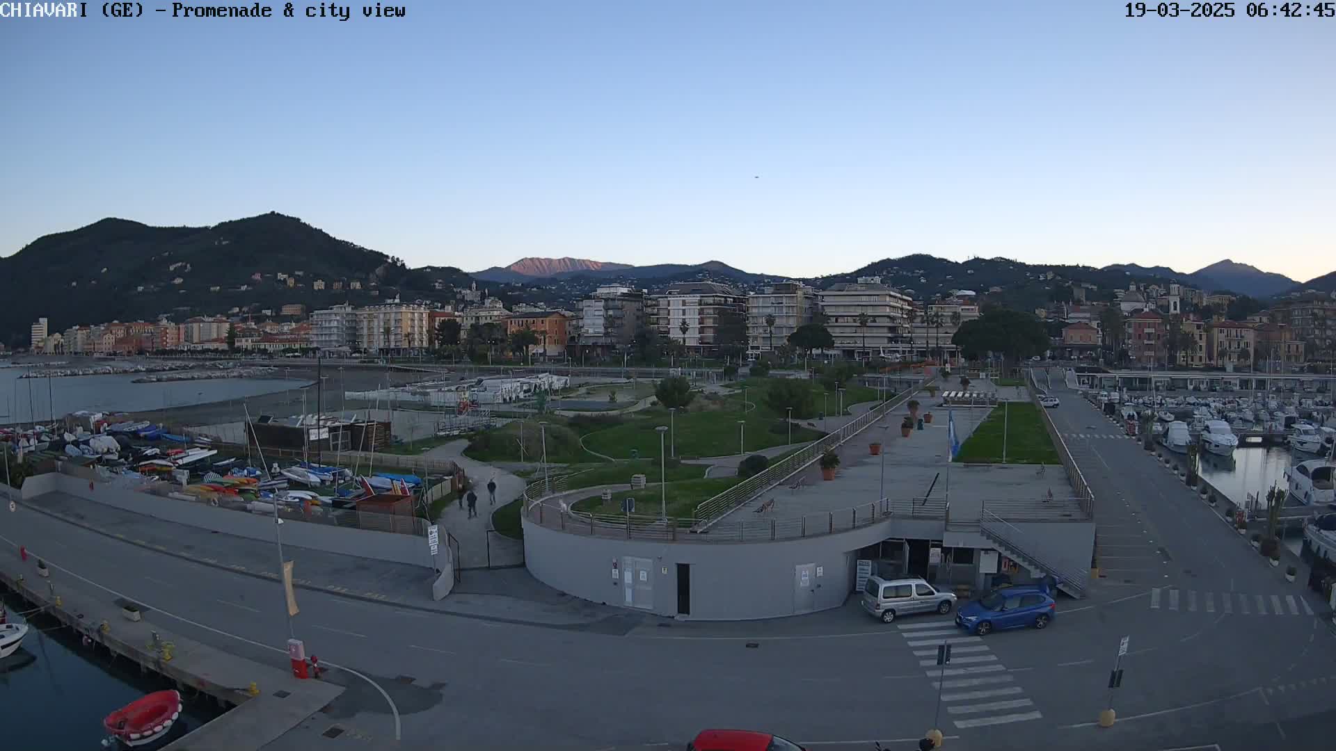 A coastal city with calm waters, a marina filled with boats, a paved promenade, and a park under a clear twilight sky.