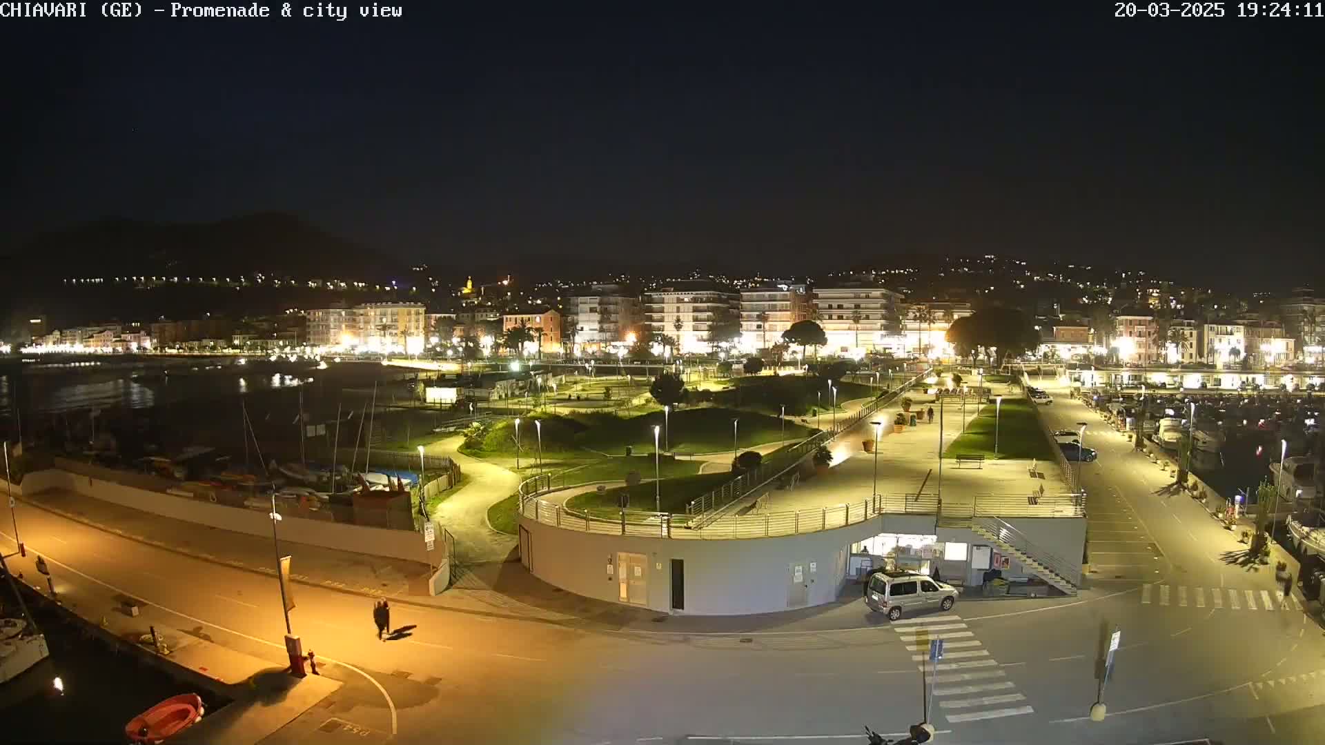 A nighttime aerial view of a harbor-side promenade and city, illuminated by streetlights, with calm water and a few parked cars.