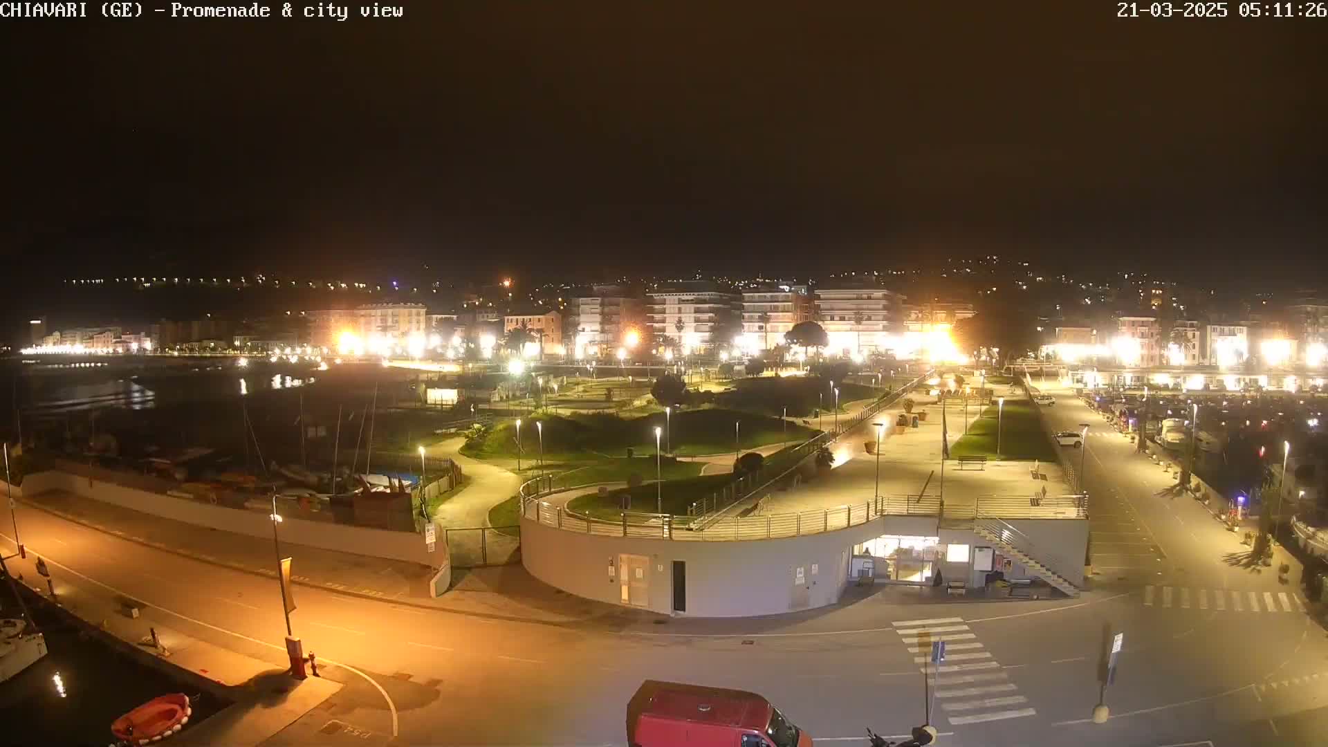 A nighttime view of a city's waterfront promenade, showing a harbor with boats, a park-like area with winding paths, and buildings lit up along the shore.