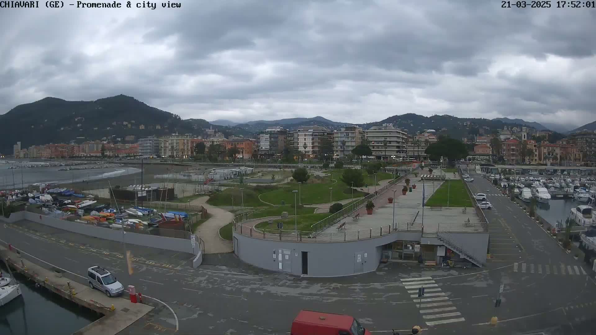 An overcast day shows a city waterfront with a marina, park, and buildings, all backed by hills.