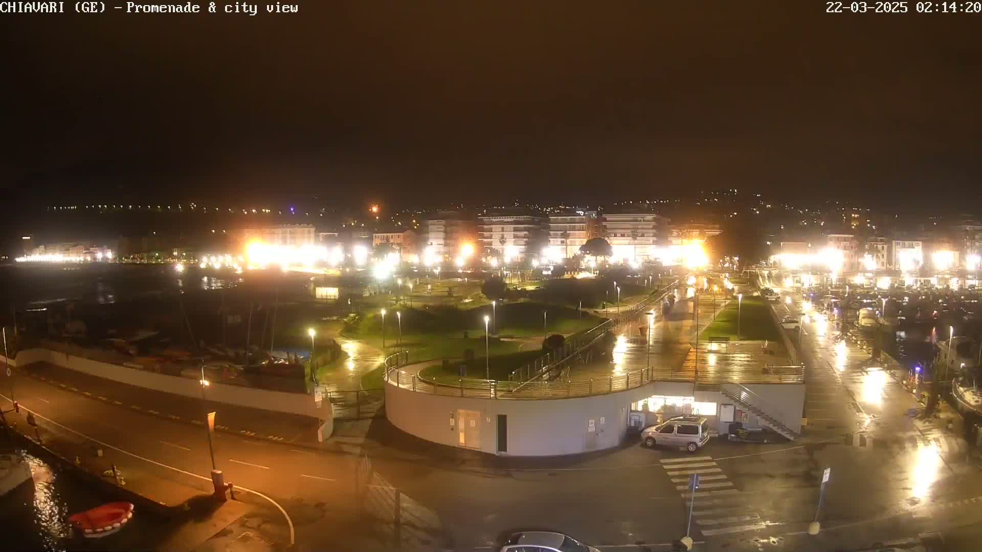 A rain-slicked nighttime view of a seaside town's promenade and harbor, illuminated by streetlights and building lights.