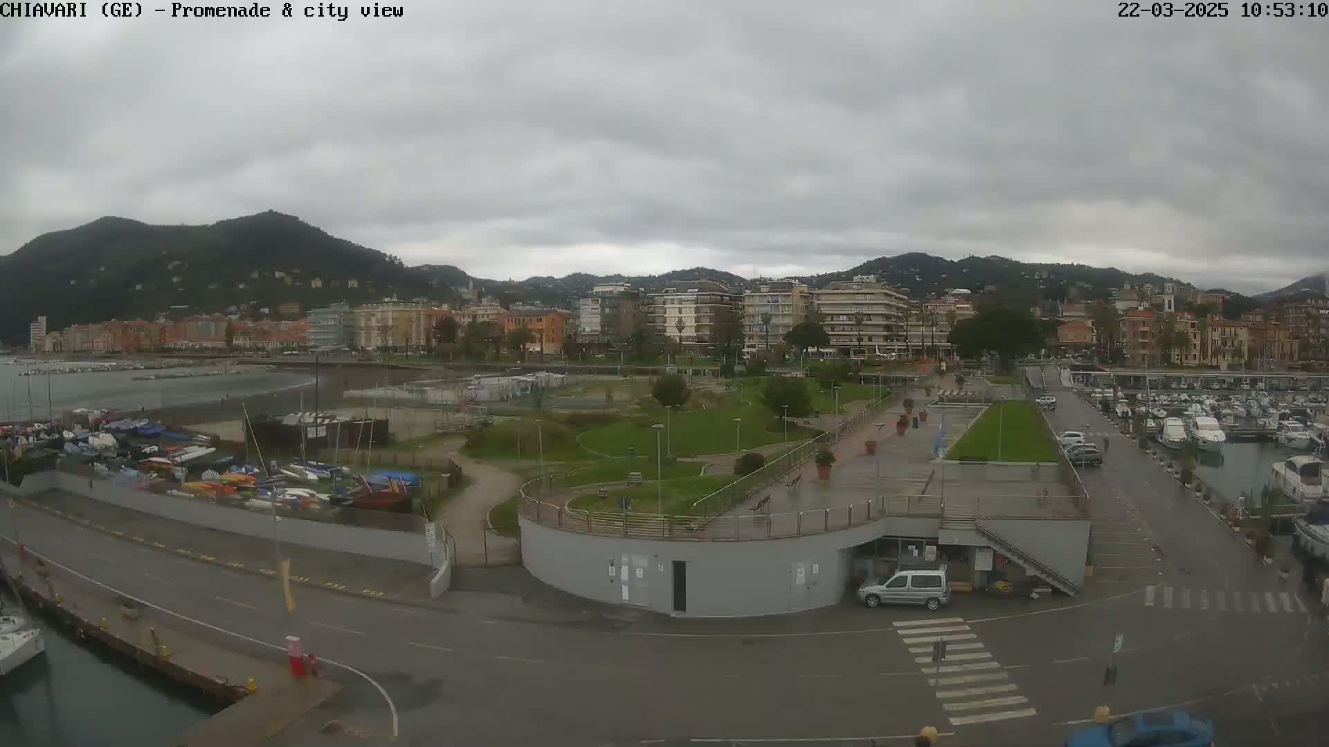 An overcast day reveals a coastal city with a promenade, a marina filled with boats, and a green park area situated between the water and the buildings.