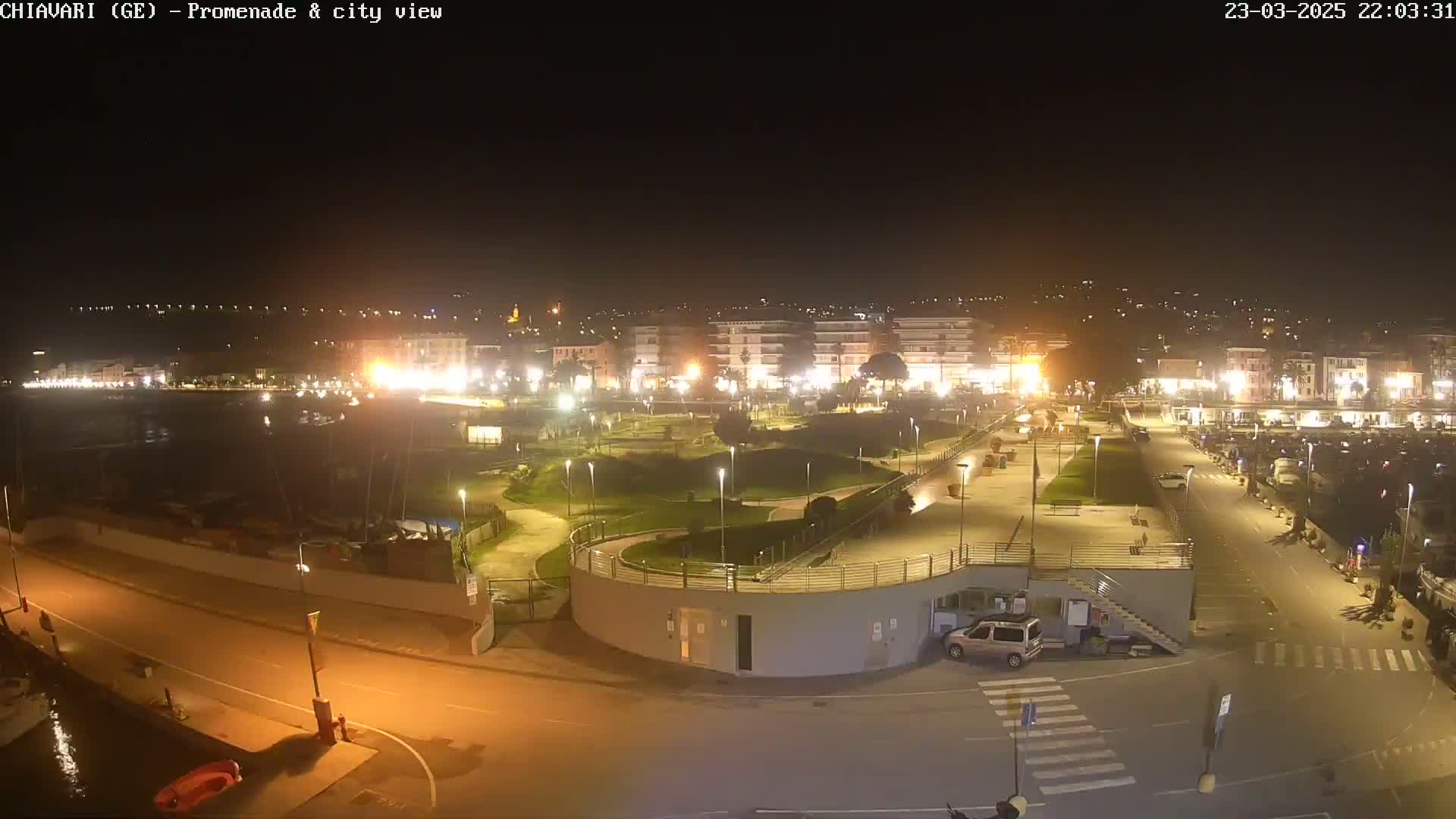 A nighttime, brightly lit waterfront promenade and city view, showing a park, buildings, and a marina.
