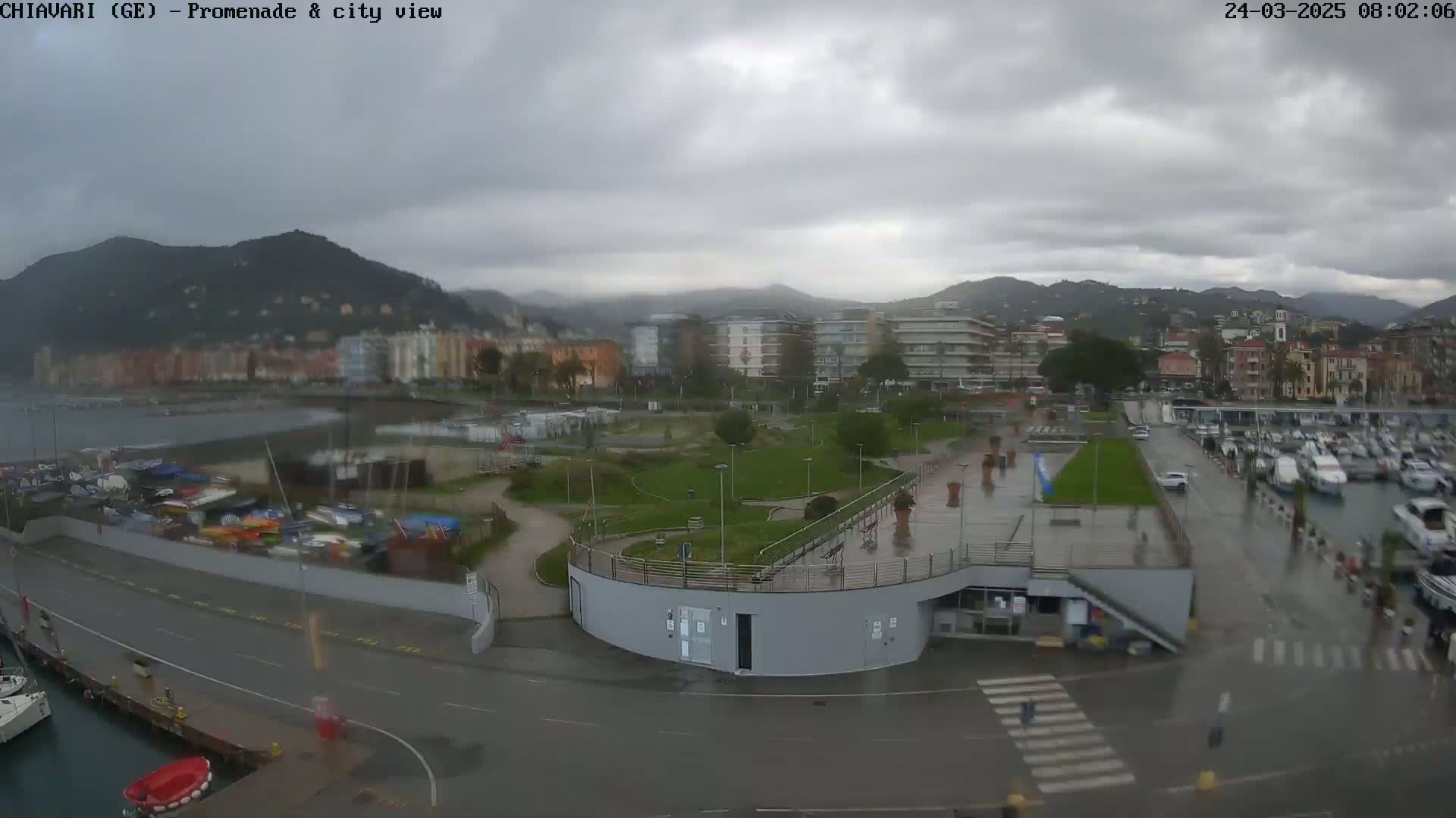 A waterfront promenade with a city and mountains in the background under an overcast sky.