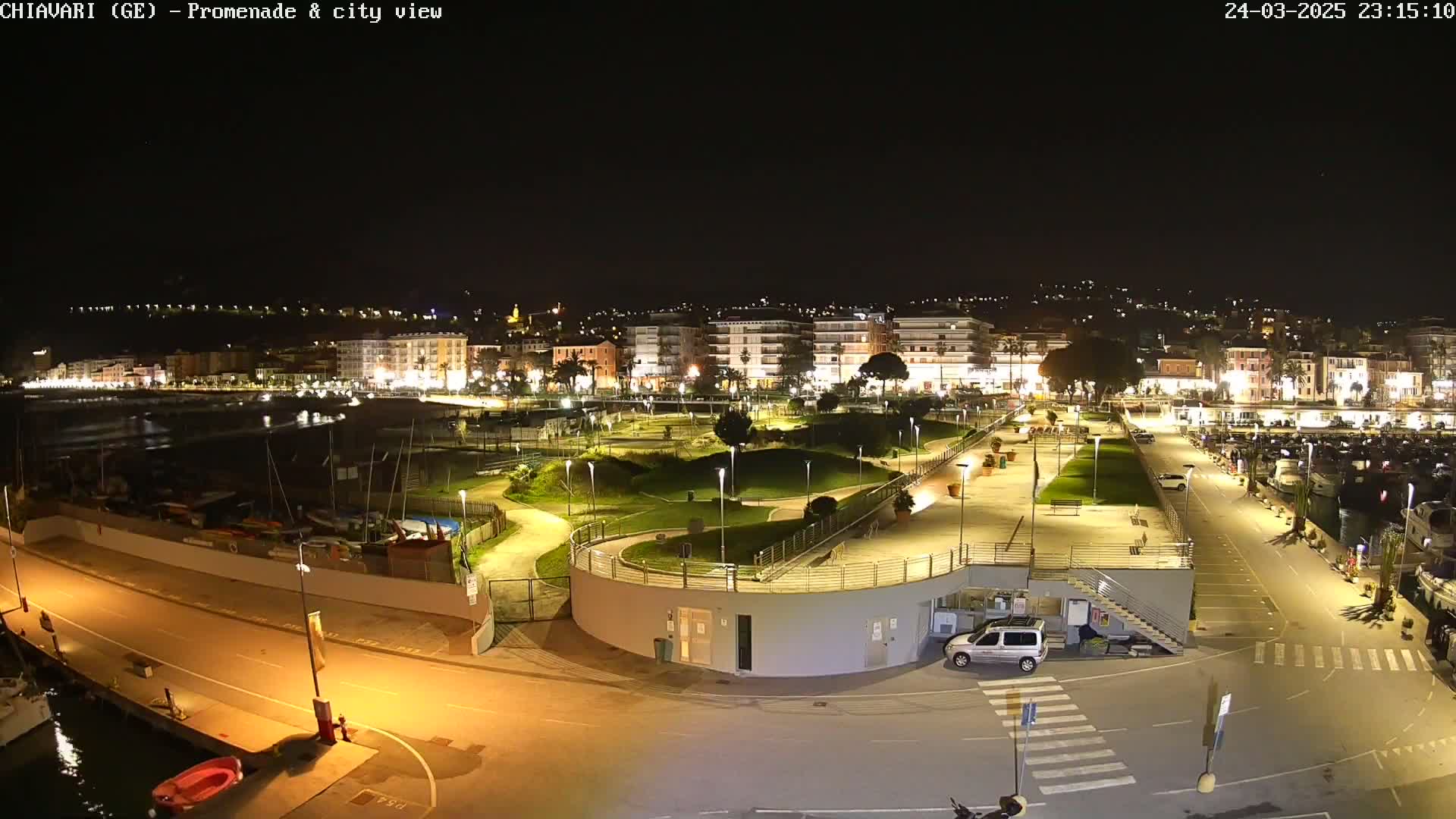 A nighttime, overhead view of a brightly lit waterfront promenade and city, featuring a marina, park, and roadways.