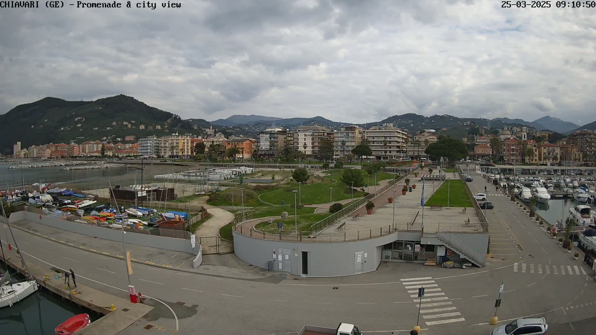 A waterfront promenade and city, under a cloudy sky, shows a harbor filled with boats, a park, and buildings extending into the distance backed by mountains.