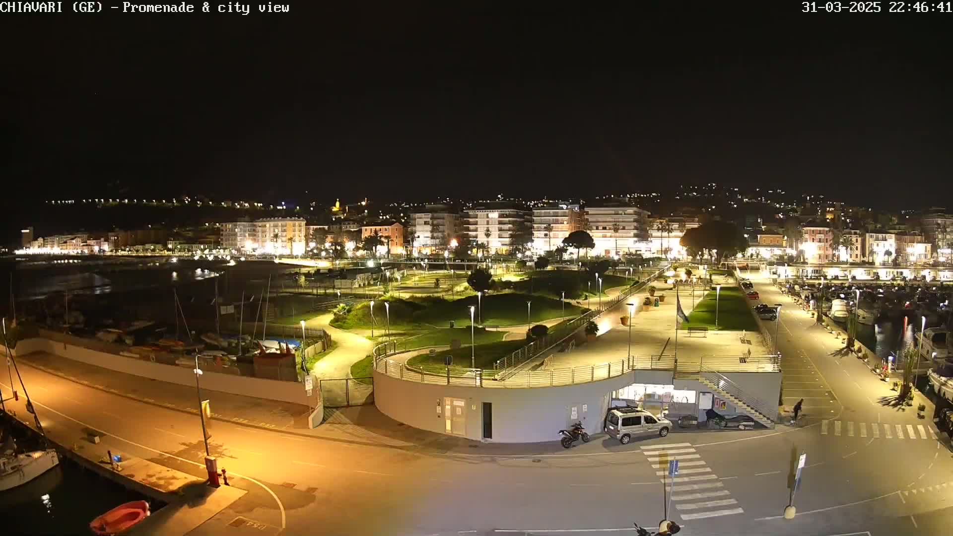A nighttime aerial view of a harbor town's waterfront promenade and marina, featuring a park with winding pathways and illuminated buildings along the shoreline.