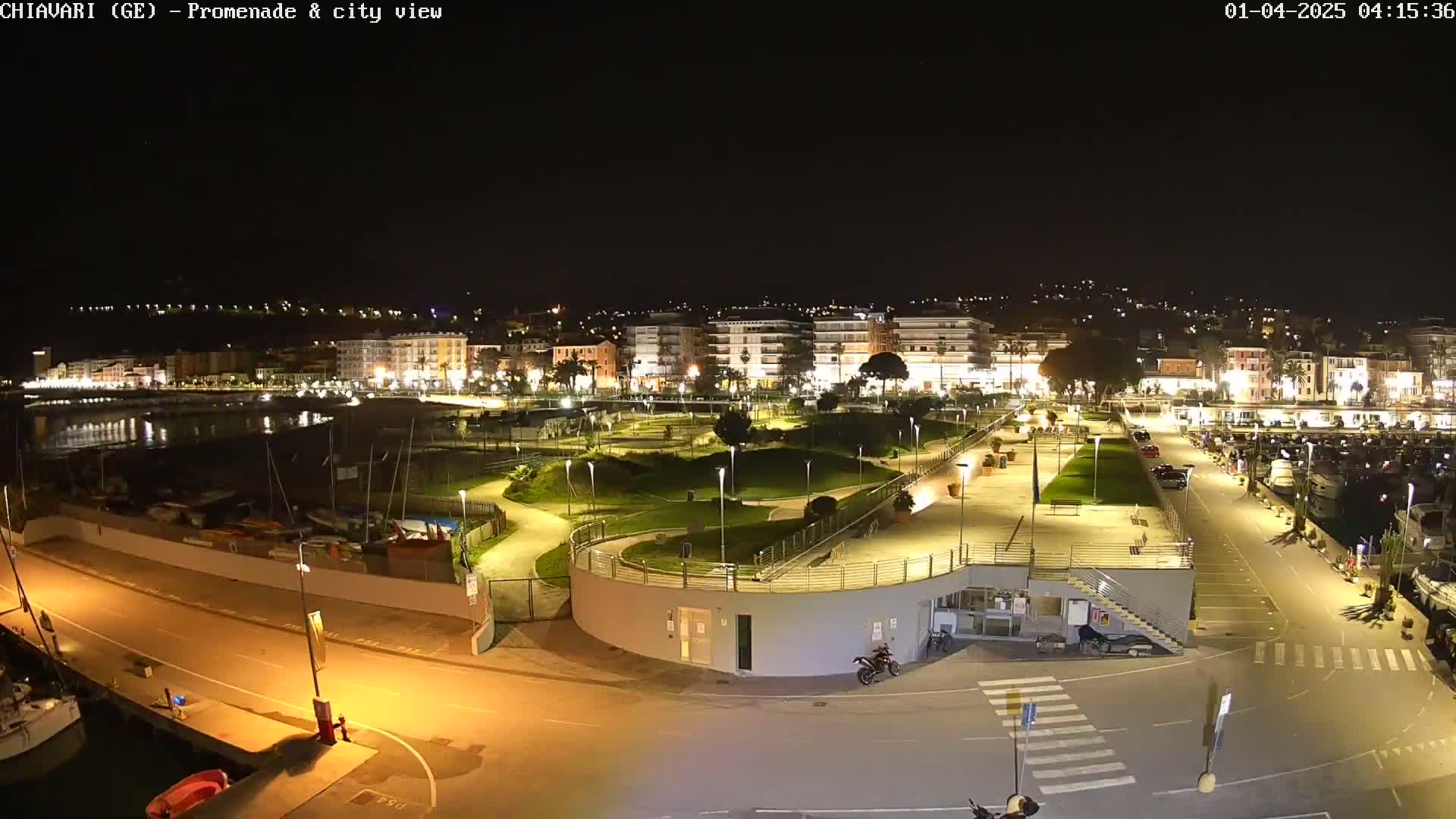 A nighttime aerial view of a waterfront promenade and city, illuminated by streetlights, showing a harbor with boats and a park-like area with winding paths.
