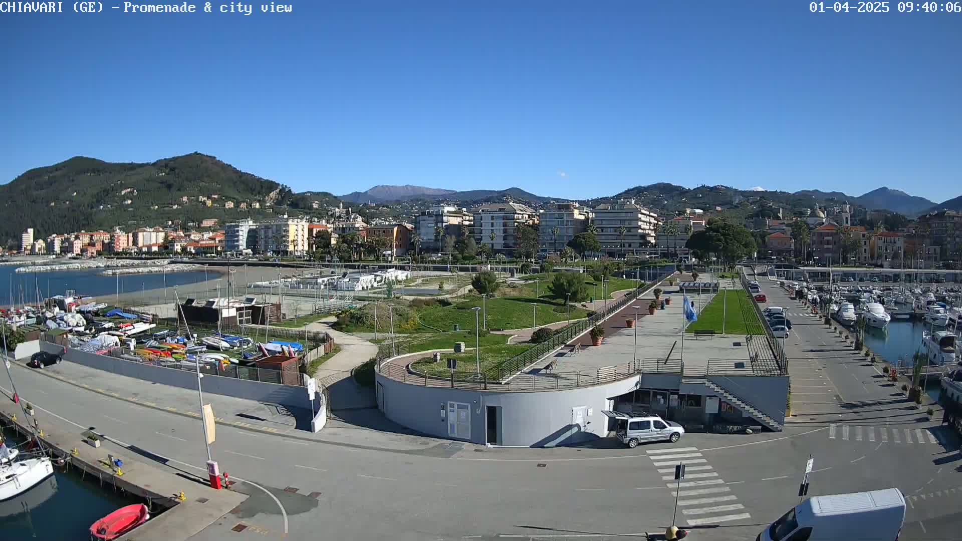 A coastal city with a marina, a promenade, and green parkland is seen on a clear, sunny day.