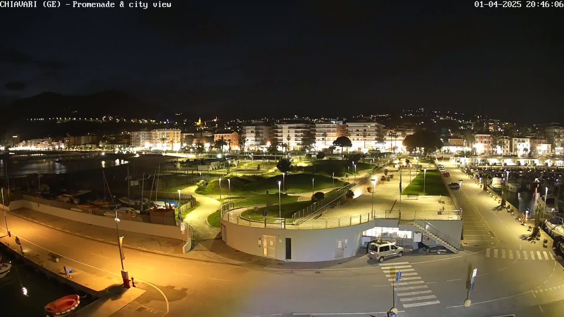 A nighttime aerial view of a city's waterfront promenade and marina, featuring a well-lit park and a small number of parked cars.