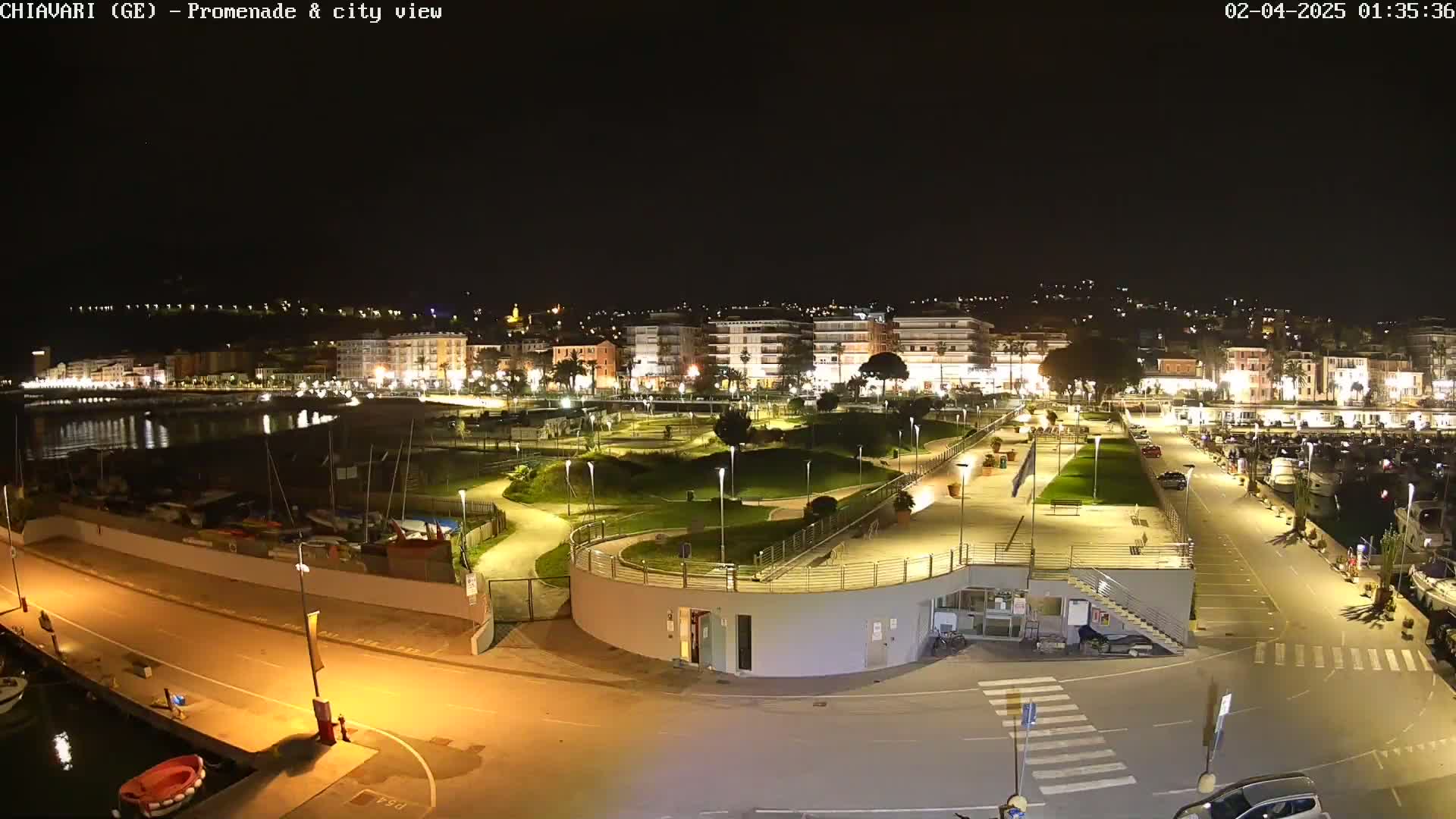 A nighttime view of a city's waterfront promenade and marina, featuring a park with winding paths and buildings illuminated against a dark sky.