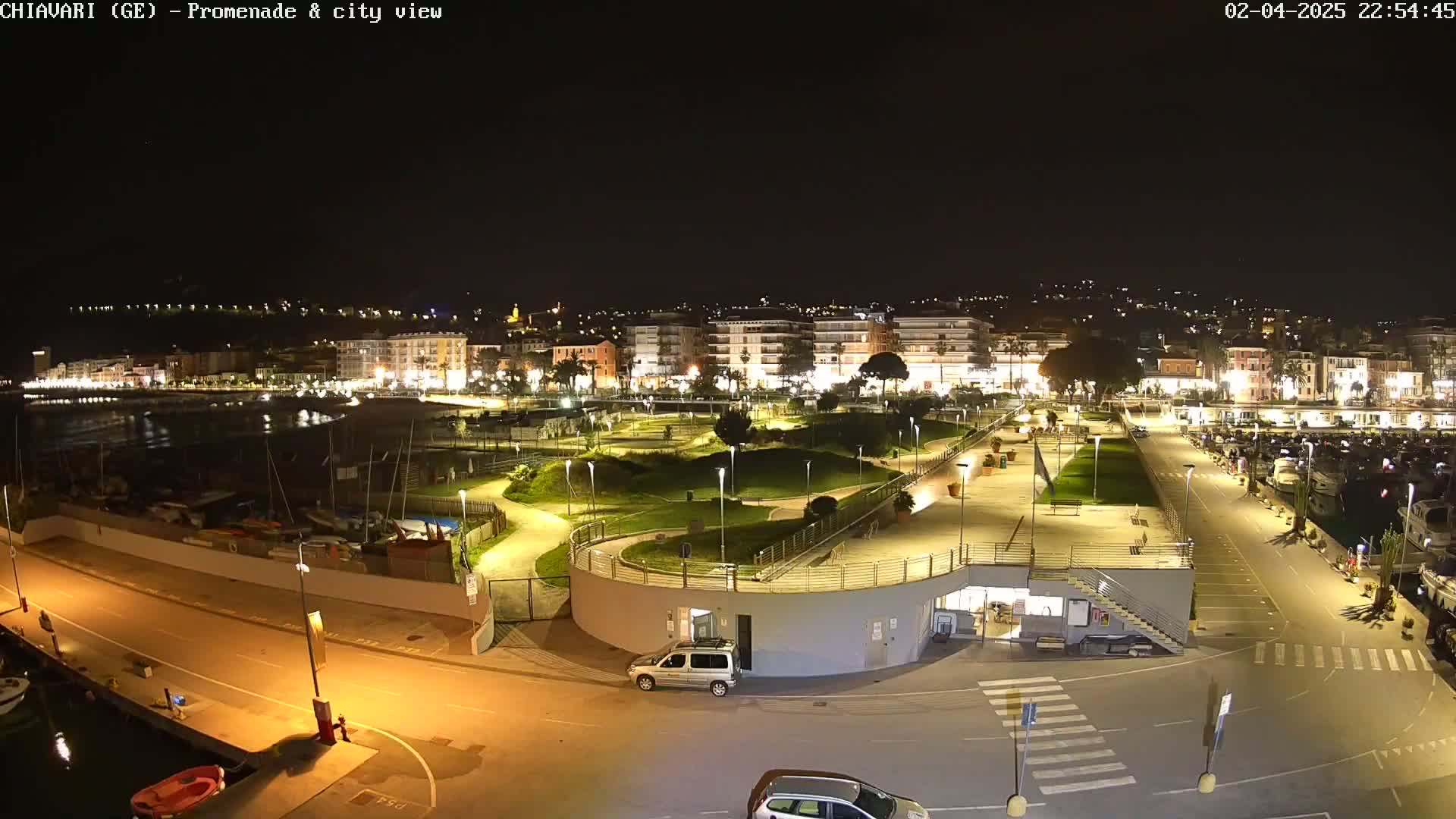 A nighttime aerial view of a city's illuminated waterfront promenade, featuring a marina, park, and buildings.