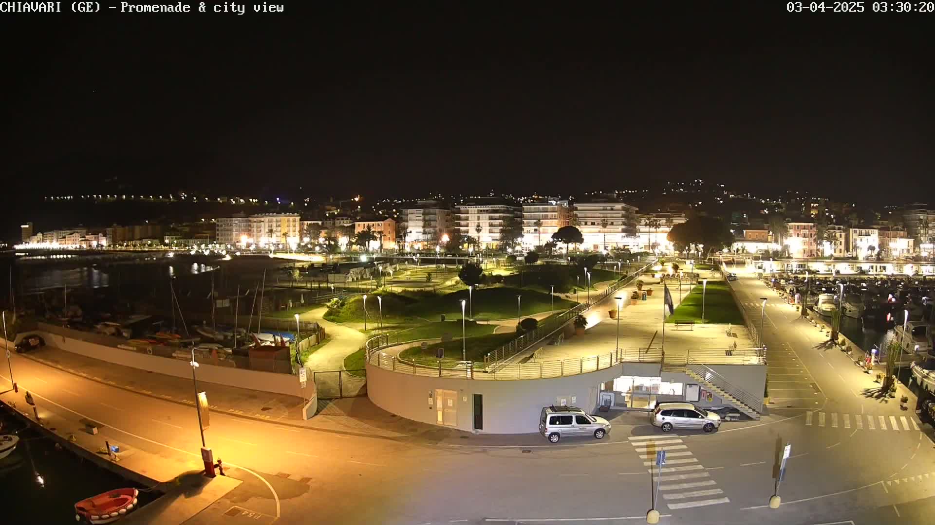 A nighttime, illuminated view of a waterfront promenade and city, featuring a harbor with moored boats, a landscaped park, and a parking lot with cars.