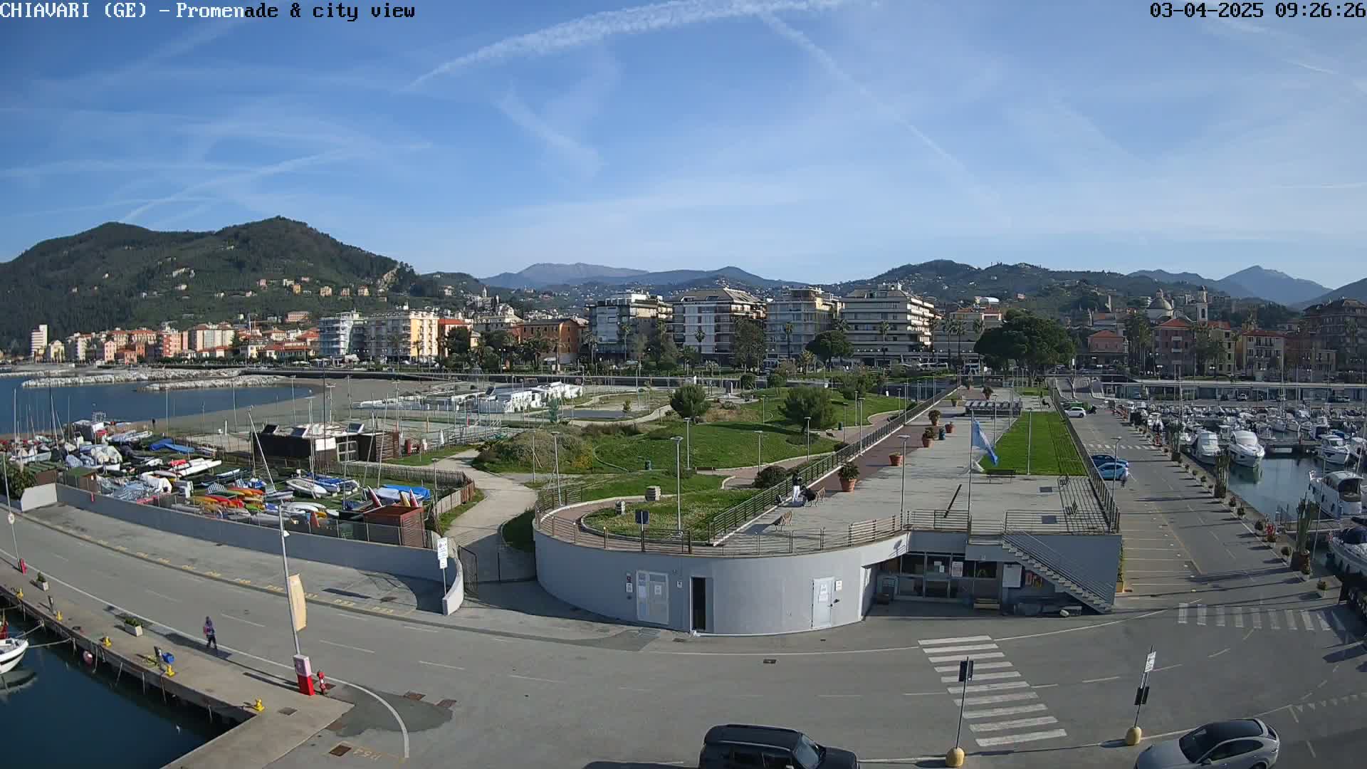 An aerial view shows a waterfront promenade with a marina filled with boats, a grassy park, and a town nestled against a mountain range under a clear, sunny sky.