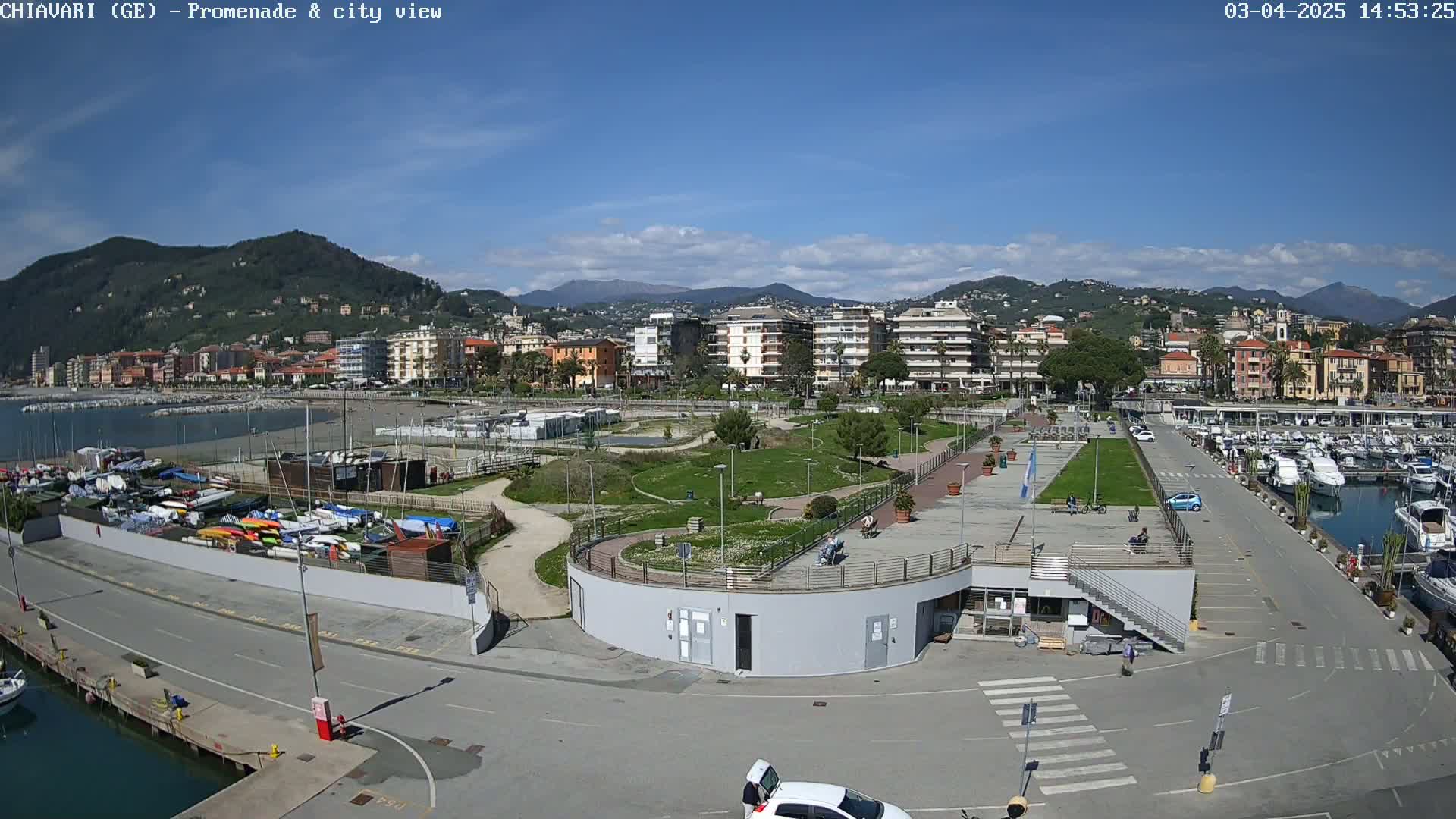 An aerial view shows a coastal town on a sunny day, with a marina filled with boats, a promenade with a park, and buildings extending up a hillside.