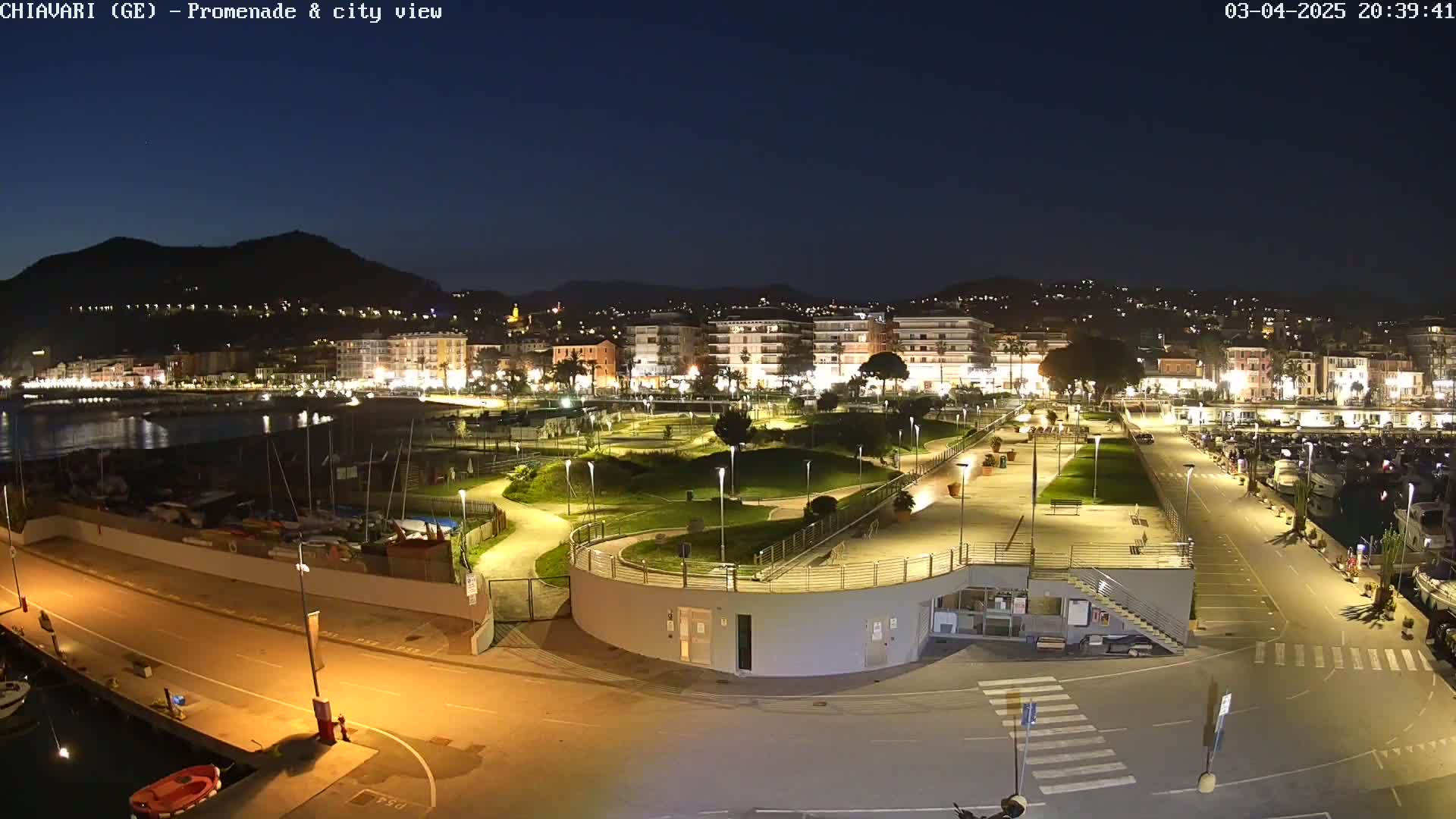 A nighttime view of a coastal city's promenade and harbor, illuminated with artificial lights, showing a park-like area with winding paths and a marina filled with boats.