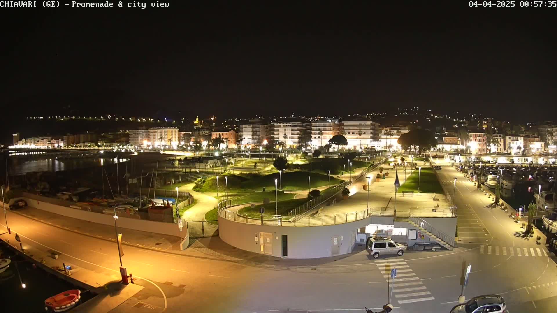 A nighttime, brightly lit waterfront promenade and city view shows a marina, park, and buildings.
