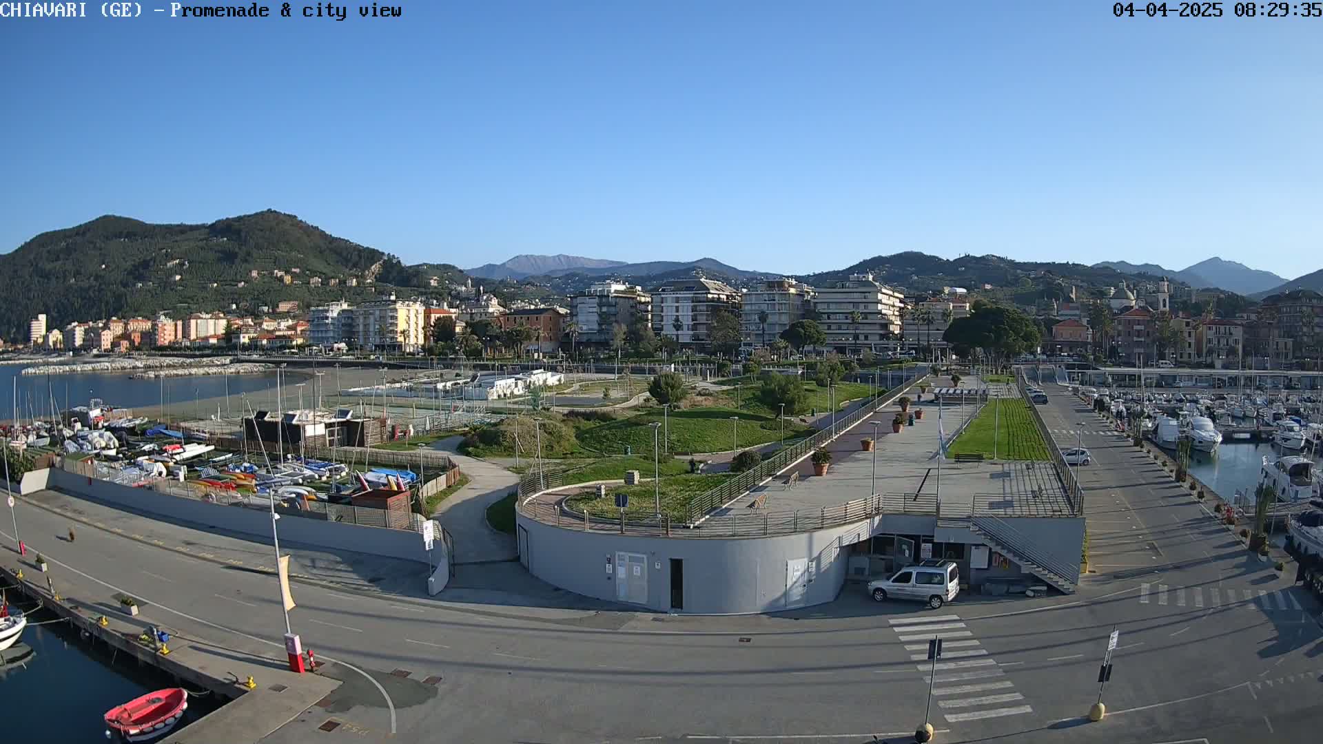 A clear, sunny day reveals a waterfront promenade with a marina full of boats, a grassy park, and a cityscape nestled against a mountain backdrop.