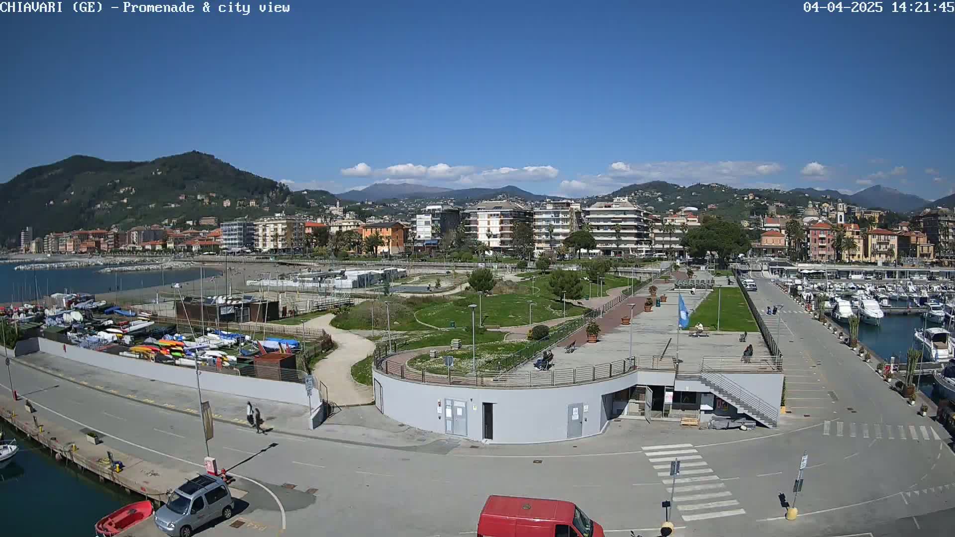 A seaside town on a sunny day with a marina filled with boats, a promenade, and a park next to a body of water, backed by hills and mountains.