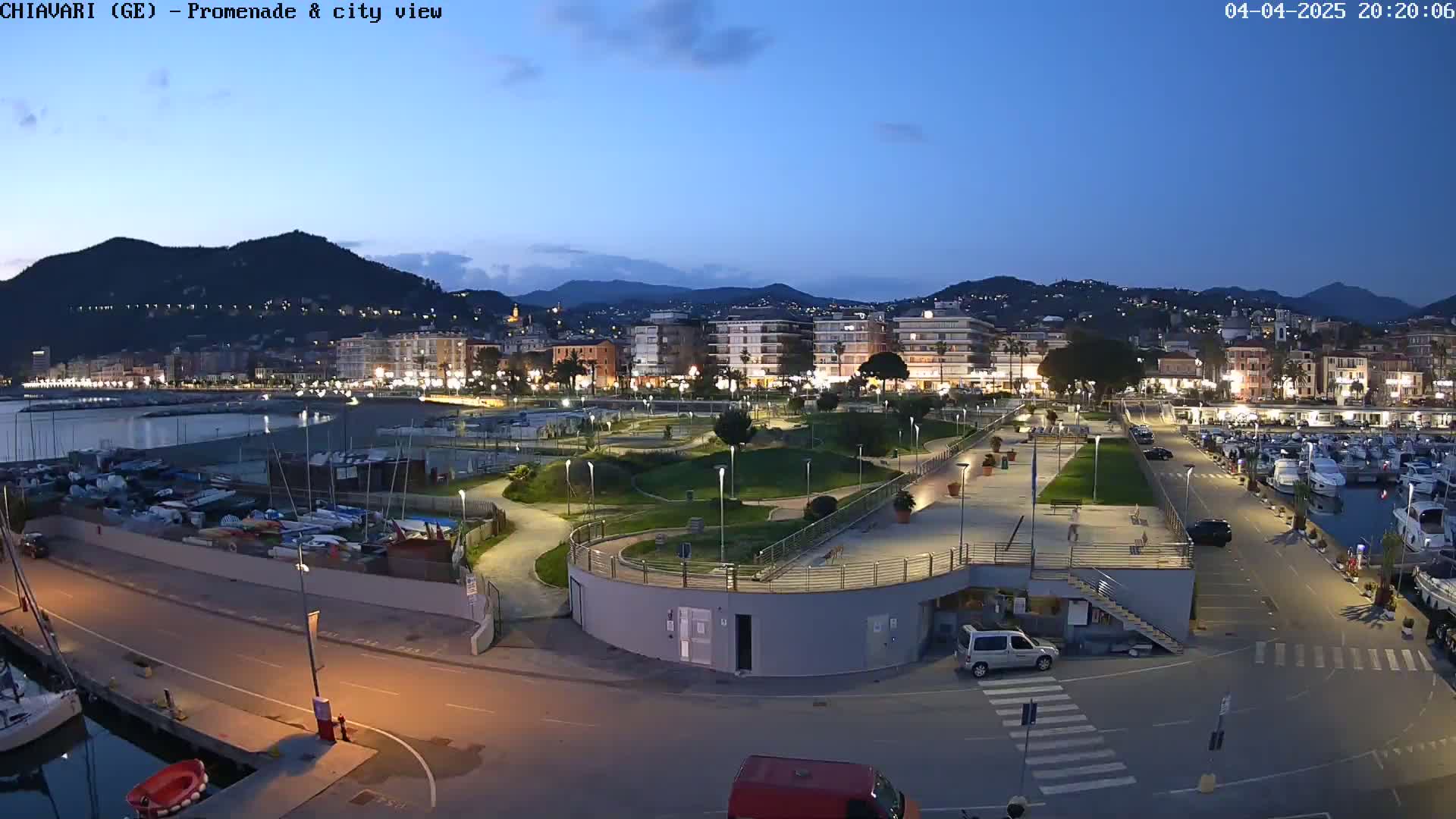 An illuminated waterfront promenade and marina at dusk, nestled between a city and hills under a clear twilight sky.