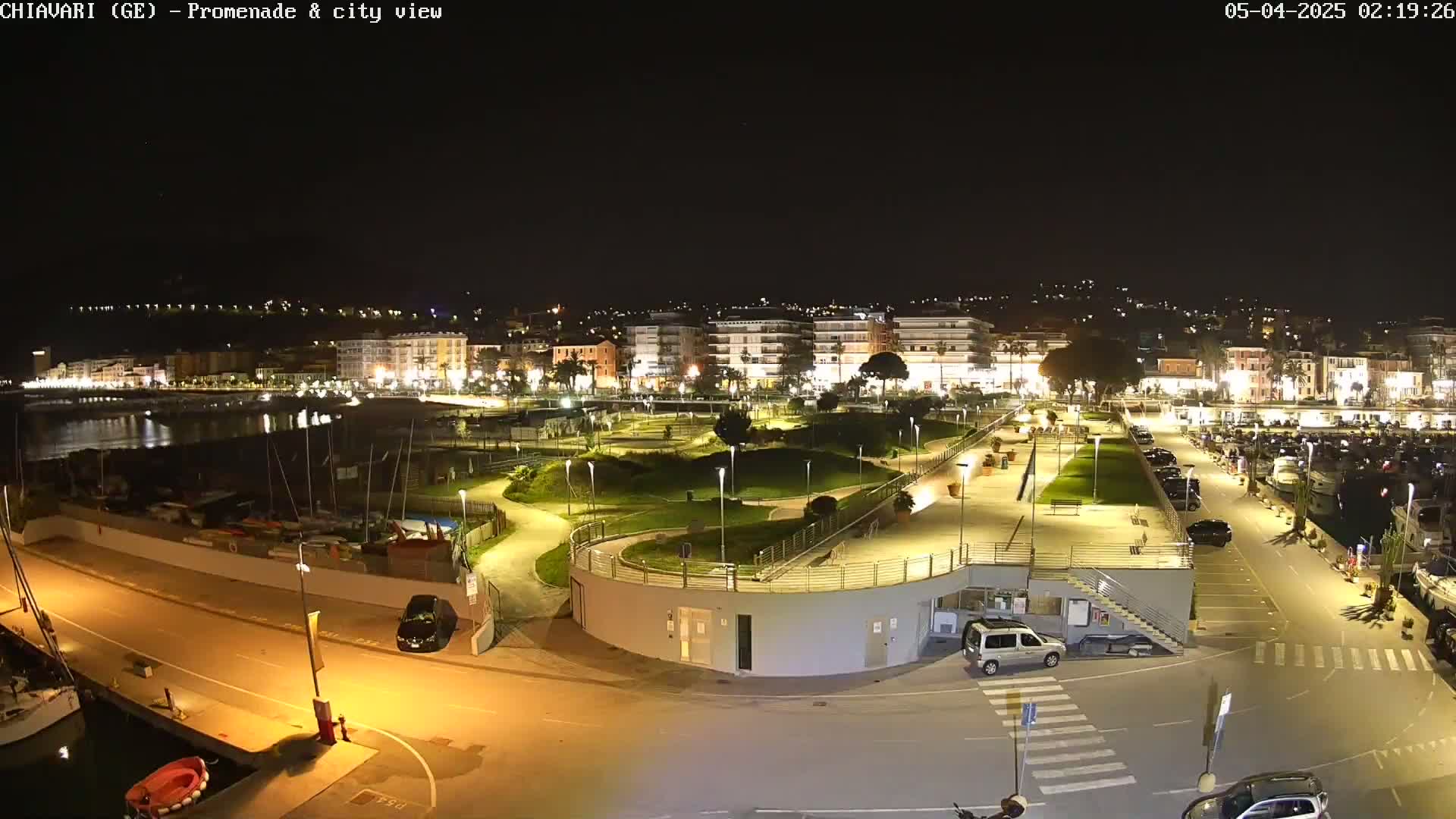 A nighttime, aerial view of a city's waterfront promenade, featuring a park, parking lot, and harbor with boats, all illuminated by artificial lights.