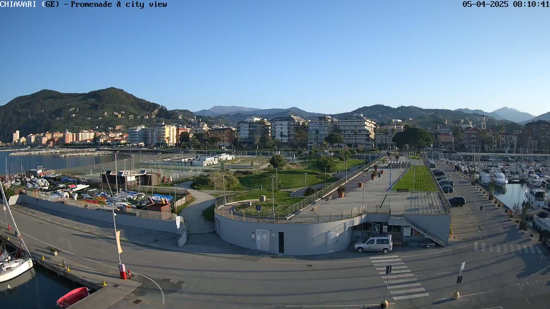A coastal town's promenade and harbor are visible under a clear, sunny sky, with boats docked and a grassy park area separating the harbor from the buildings.