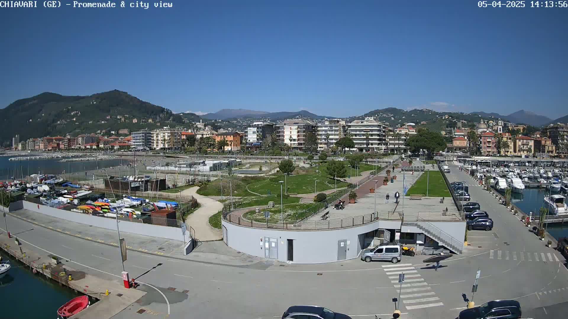 A coastal town's promenade and marina are shown on a sunny day, with a harbor full of boats and buildings along the shore backed by mountains.