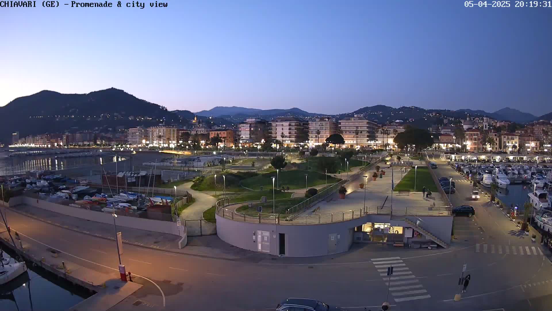 A coastal city at dusk, with a harbor full of boats and a park-like promenade, is lit by streetlights under a clear, twilight sky.