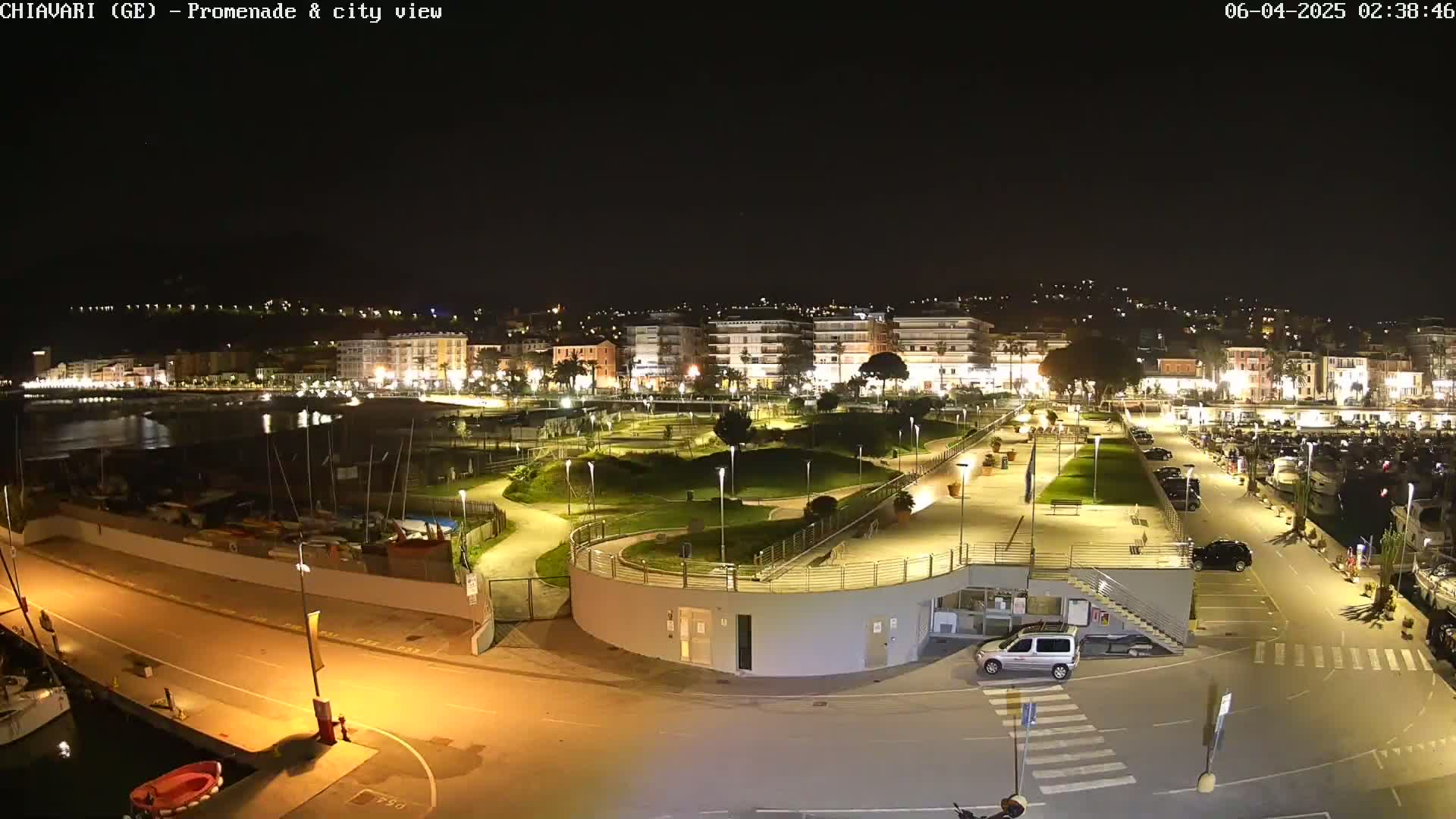 A nighttime view of a city's waterfront promenade, featuring a park, marina, and buildings illuminated under a clear, dark sky.