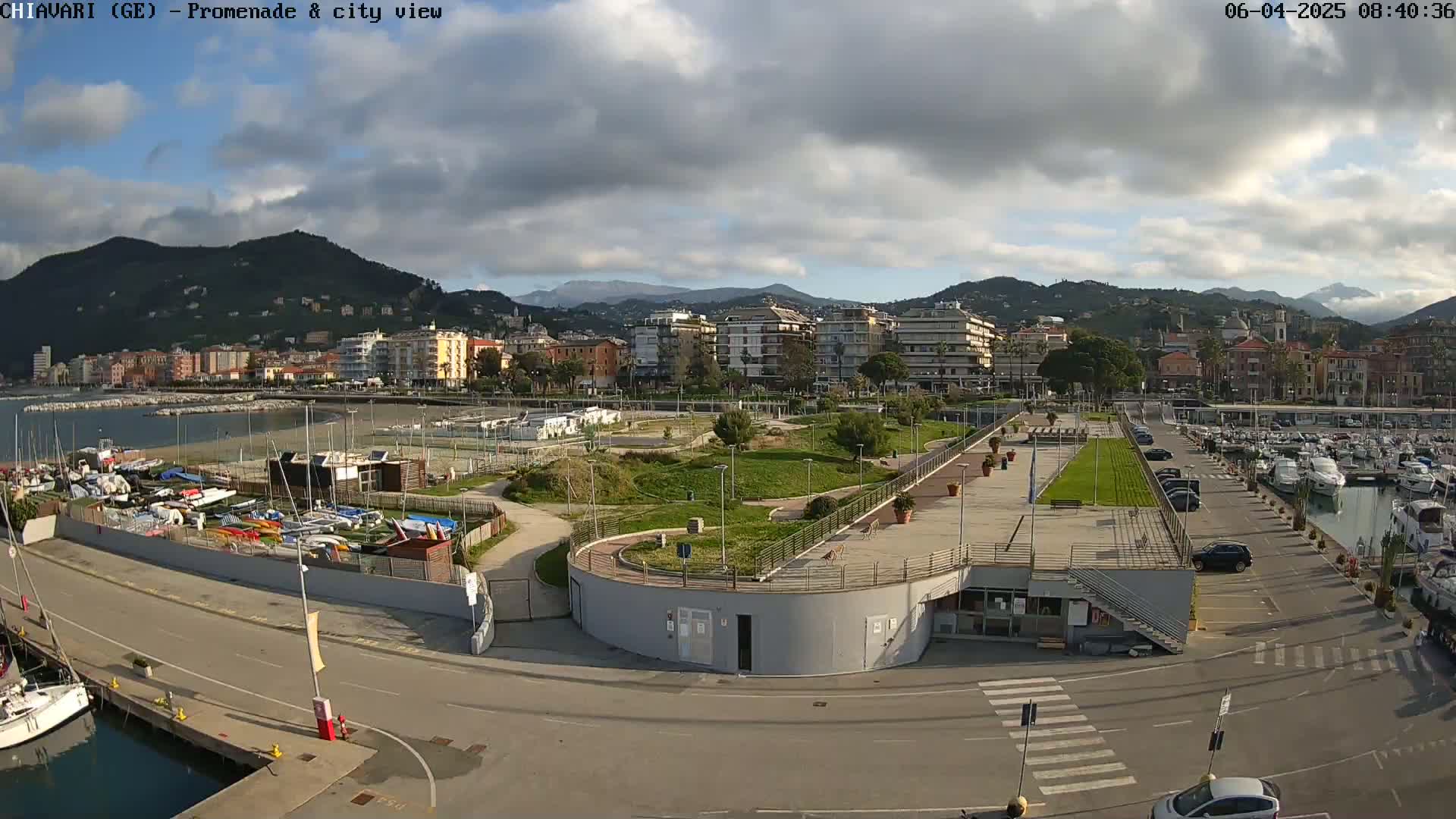 An aerial view of a coastal city with a marina, park, and buildings under a partly cloudy sky.