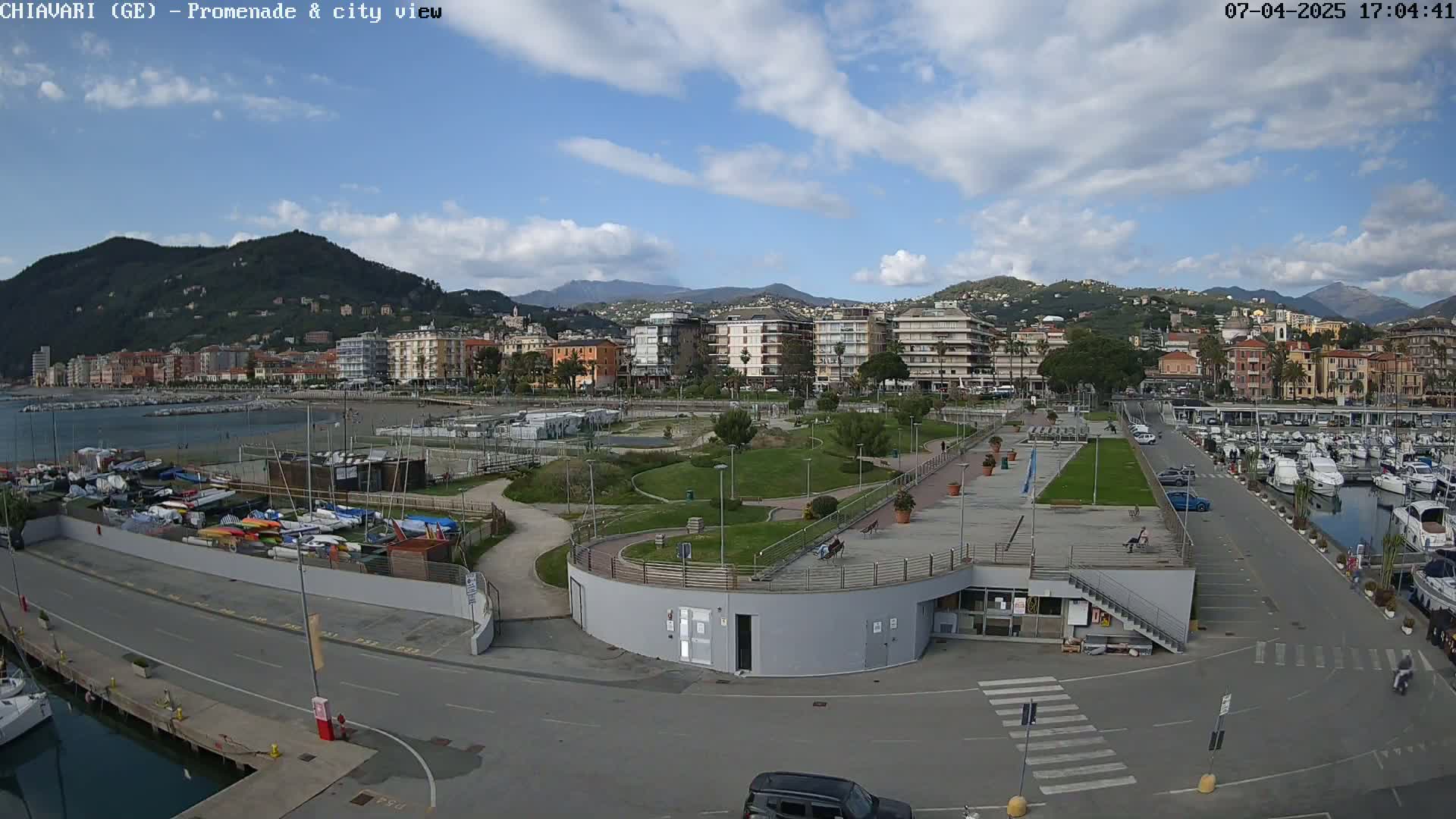 A seaside town under a partly cloudy sky shows a marina filled with boats, a paved promenade, and a green park area next to a collection of buildings extending uphill into the mountains.