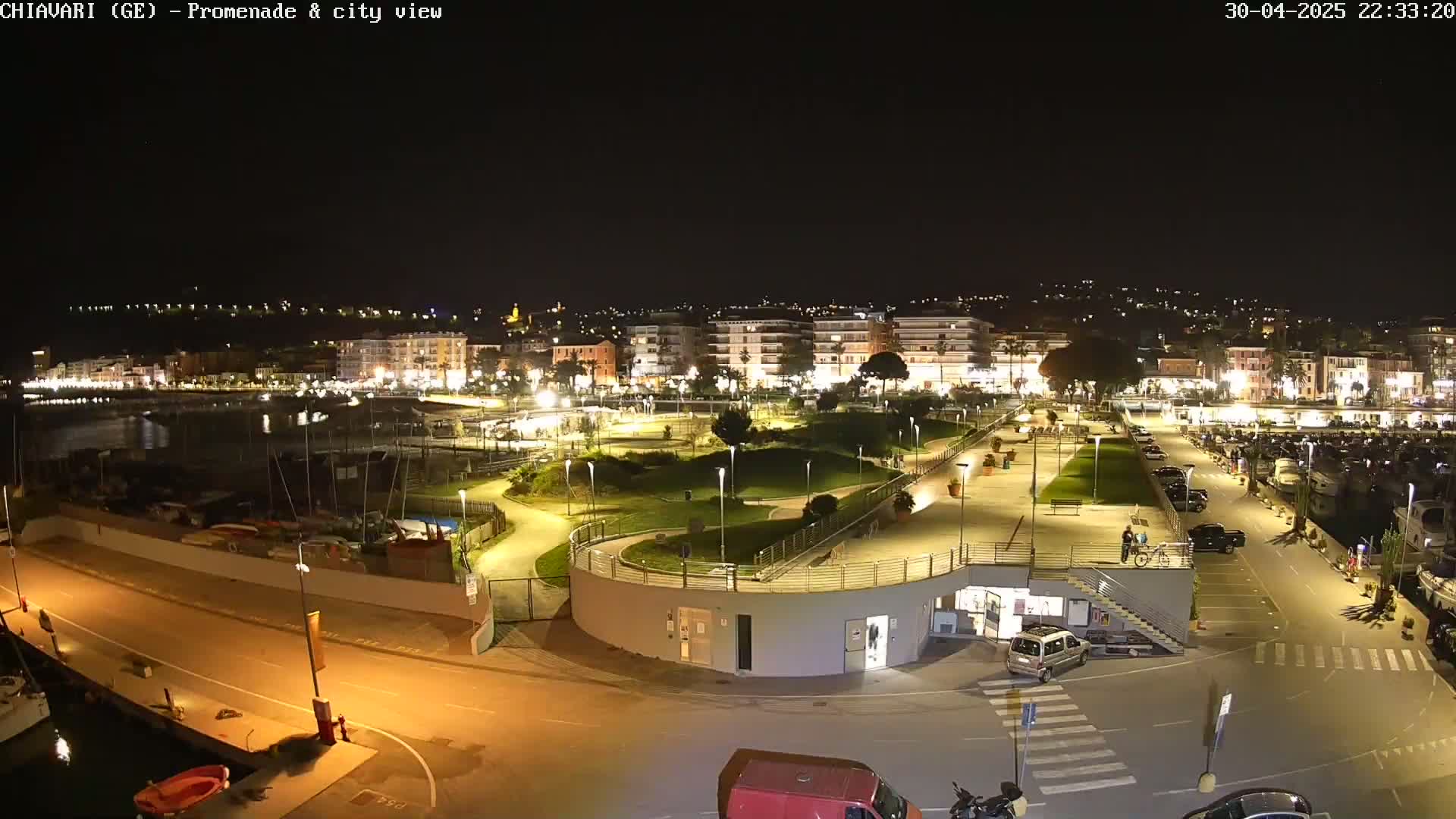 A nighttime view of a city's illuminated waterfront promenade, park, and marina, with boats and cars visible.
