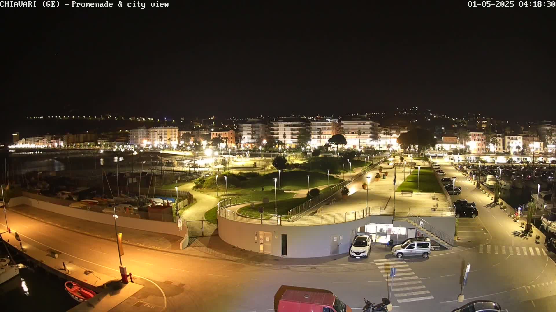 A nighttime, illuminated view of a harbor, promenade, and city, with a park separating the waterfront from the buildings.