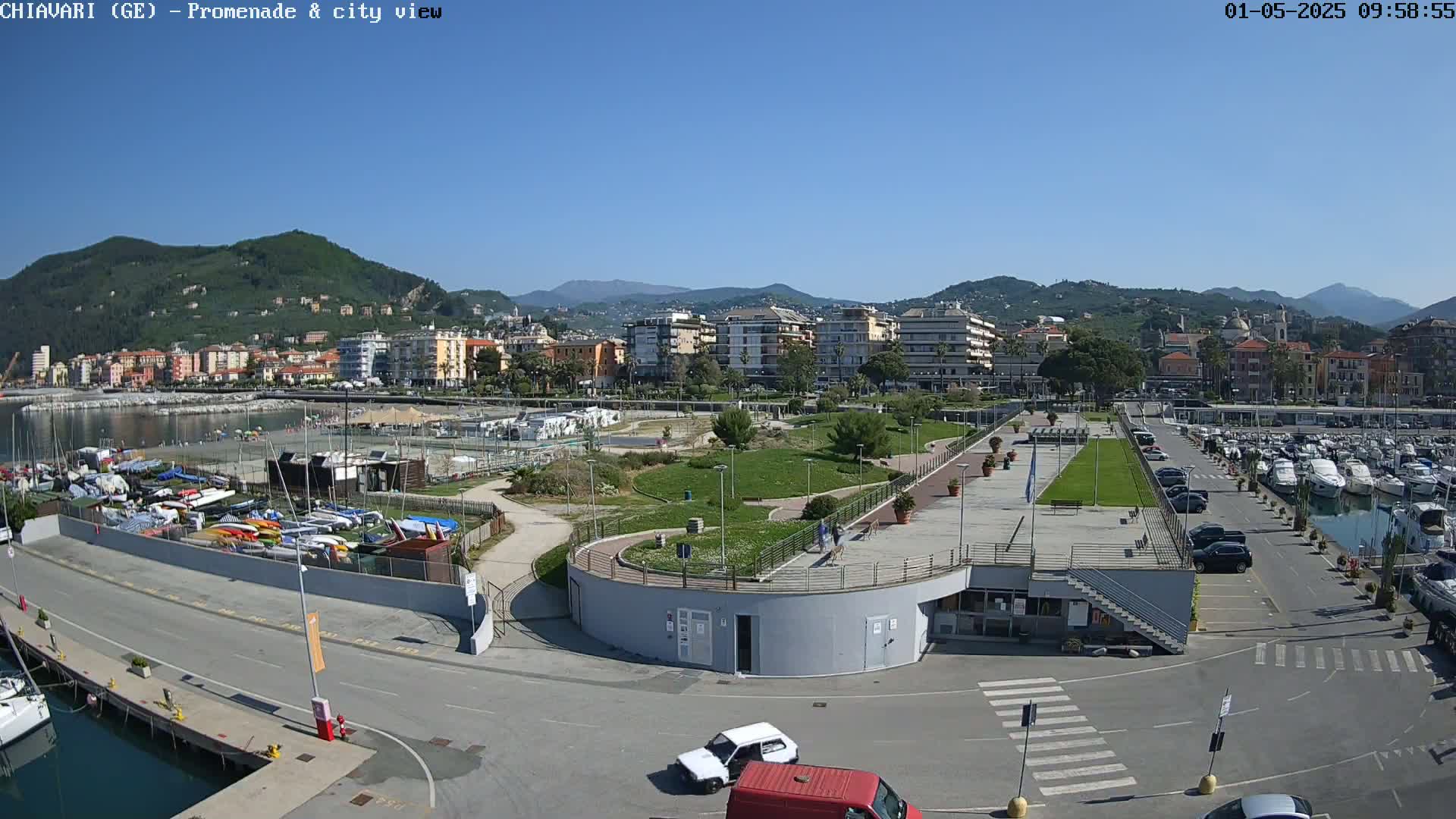 A coastal town scene on a sunny day, showing a harbor with numerous boats, a park with walking paths, and buildings nestled against a hillside.