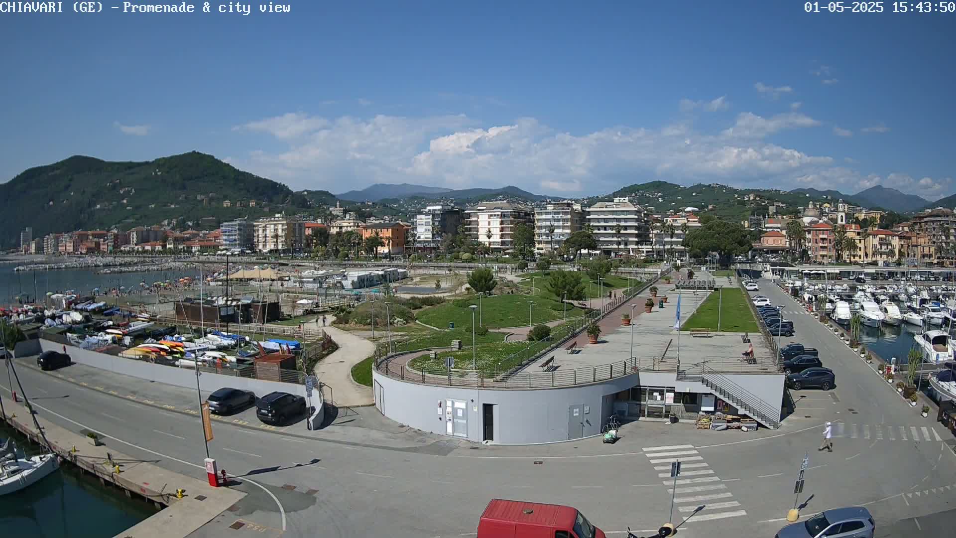 A seaside town on a sunny day with a partially cloudy sky, featuring a marina, promenade, park, and buildings nestled against a mountain backdrop.