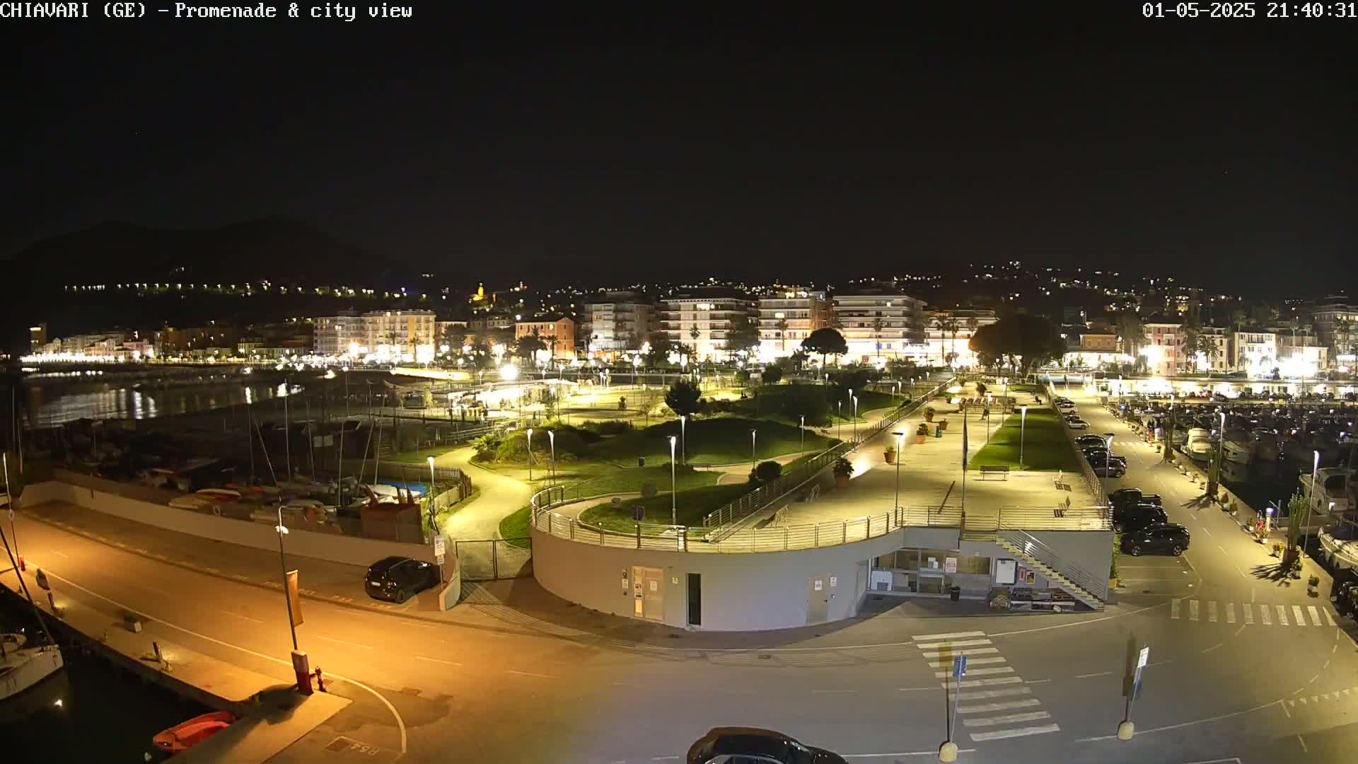 A nighttime aerial view shows a harbor with a city illuminated behind it,  a promenade and park with winding paths and landscaping in the foreground.