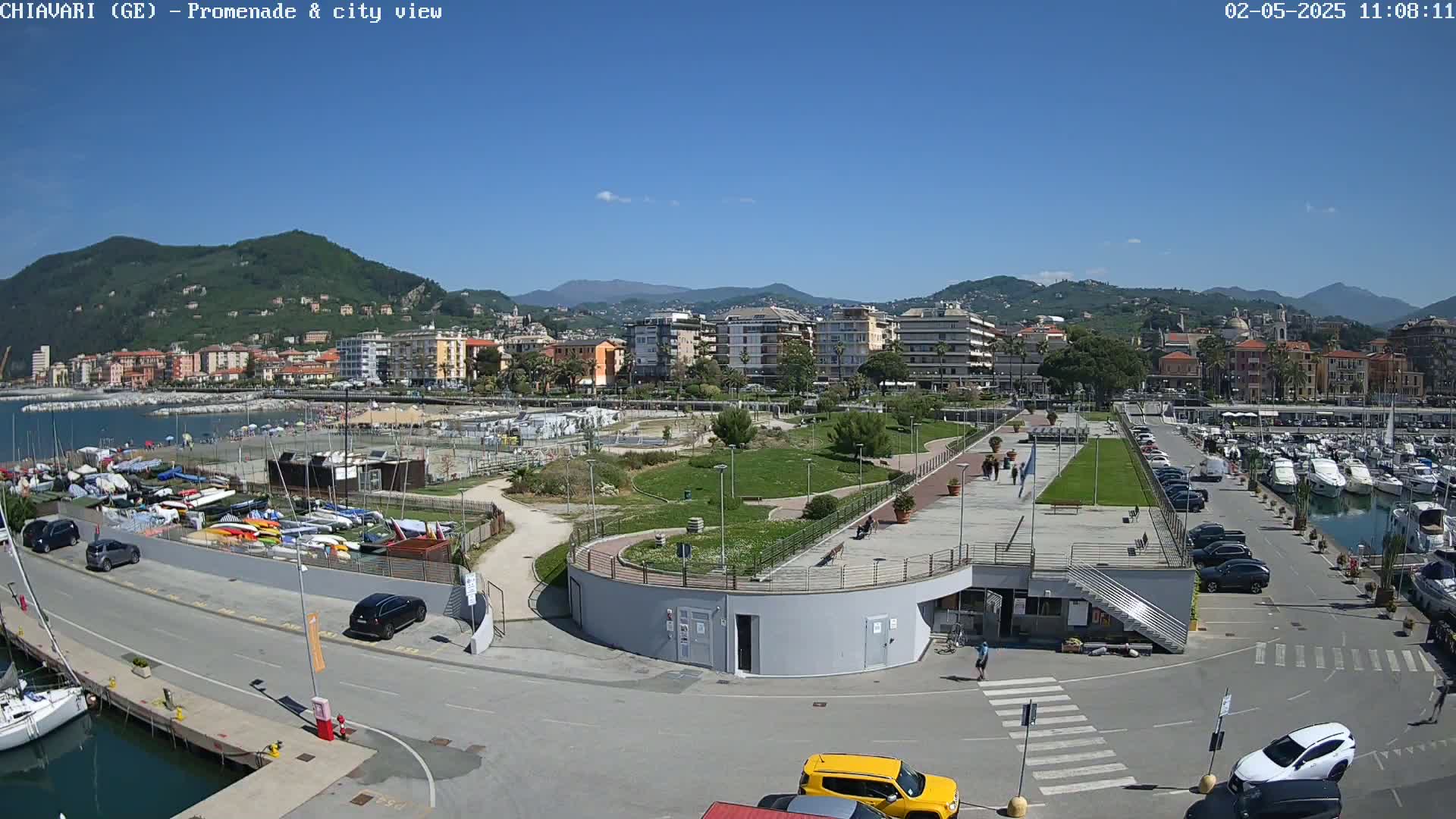 A sunny coastal town scene shows a harbor filled with boats, a promenade with a grassy park, and buildings nestled against hills in the background.
