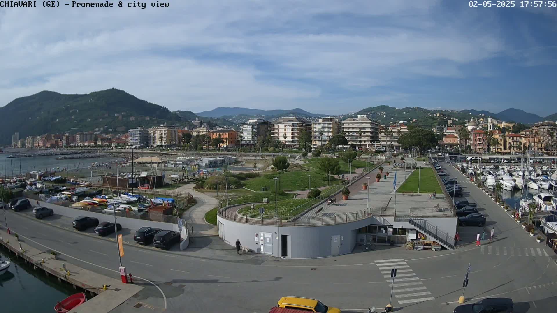 An aerial view shows a waterfront promenade with a marina full of boats, a park, and a city nestled against a mountain range under a mostly sunny sky.