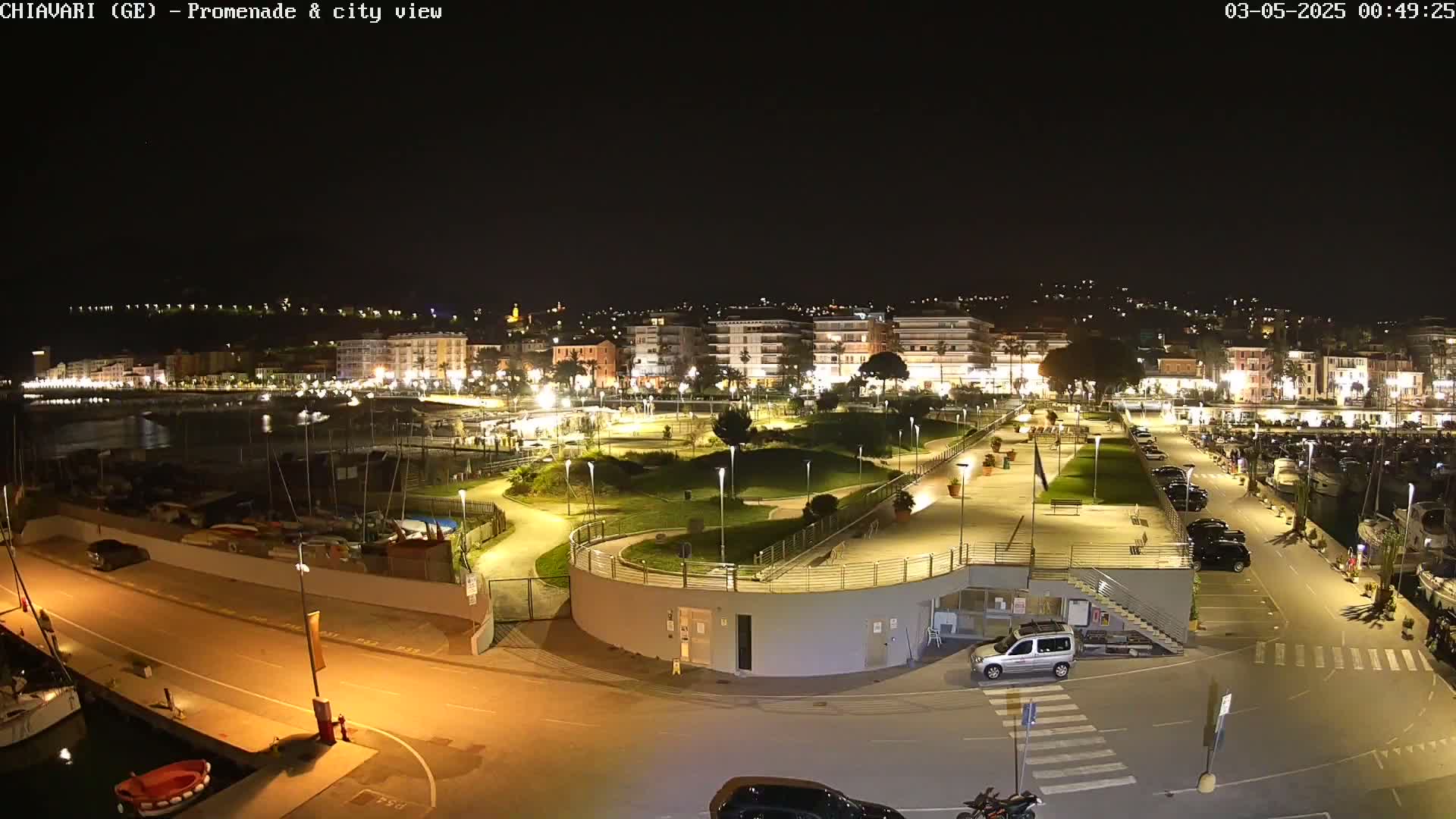 A nighttime, brightly lit waterfront promenade and city scene with a marina filled with boats, and cars parked in lots along the walkway.