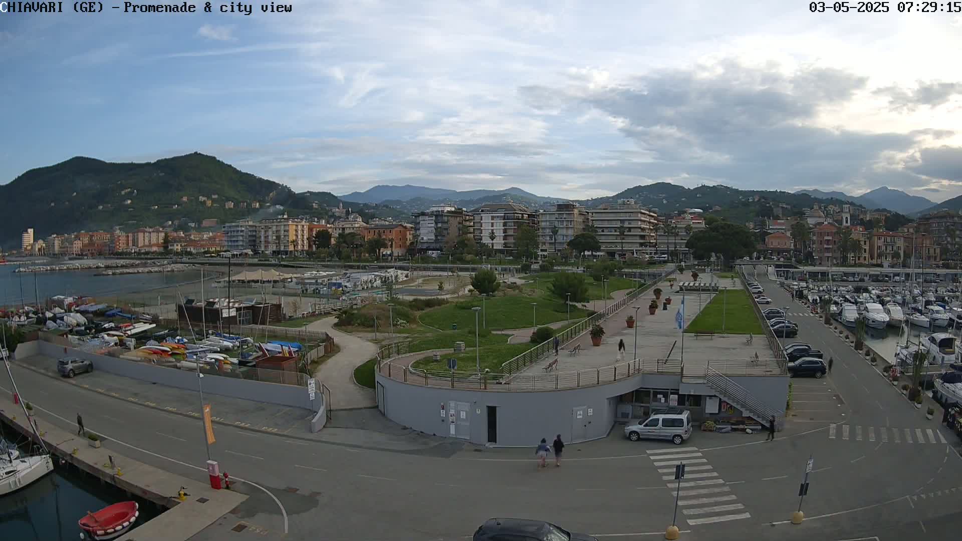 An aerial view shows a waterfront promenade with a marina, a park, and a town nestled against a mountain range under a mostly cloudy sky.