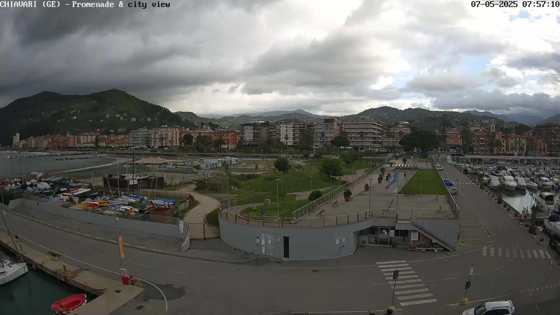 An overcast day reveals a coastal city scene with a marina filled with boats, a promenade, and green spaces, all nestled against a backdrop of mountains.